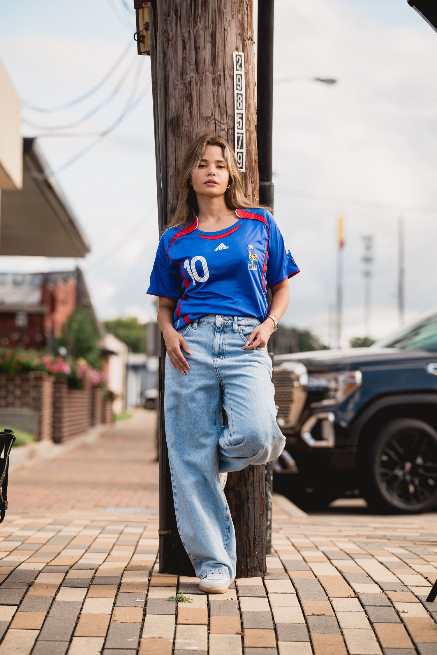 Person in blue sports jersey and jeans posing against brick wall in urban setting.