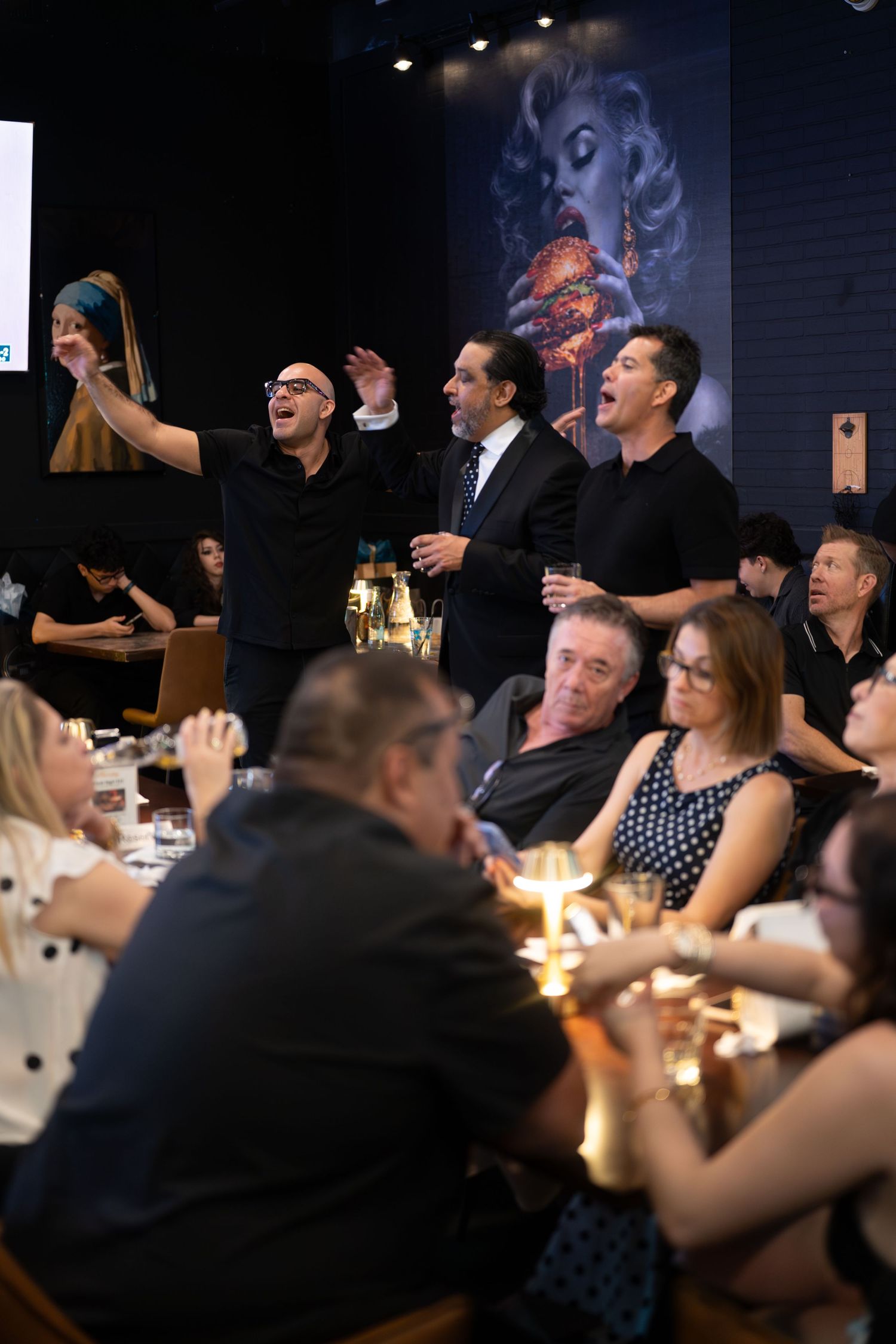 Three men stand and celebrate at a lively restaurant dinner event with guests seated at candlelit tables.