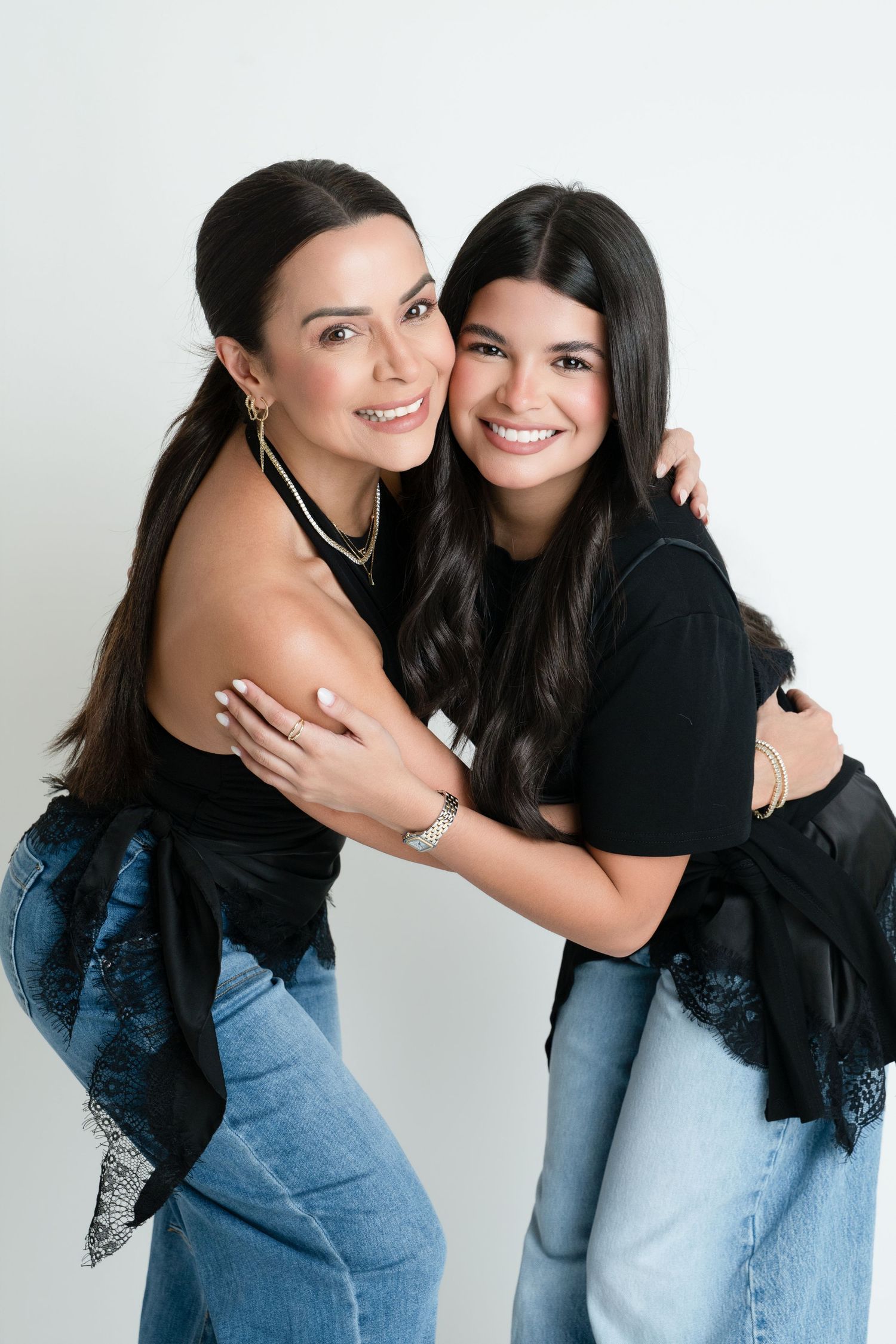Two smiling brunette women in black tops and jeans embrace warmly against a white background.