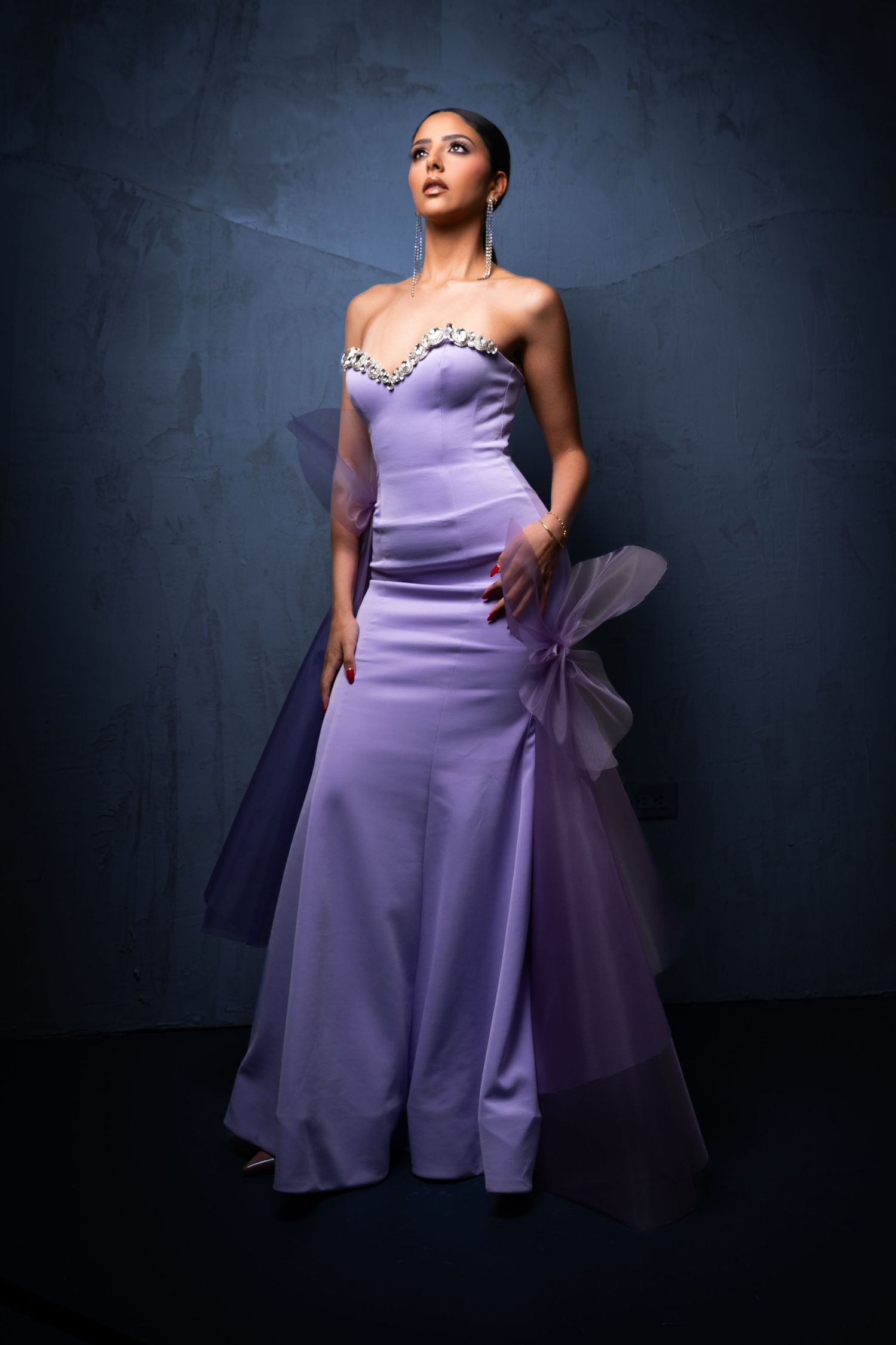Model in flowing lavender evening gown poses dramatically against dark backdrop in artistic fashion photo series.