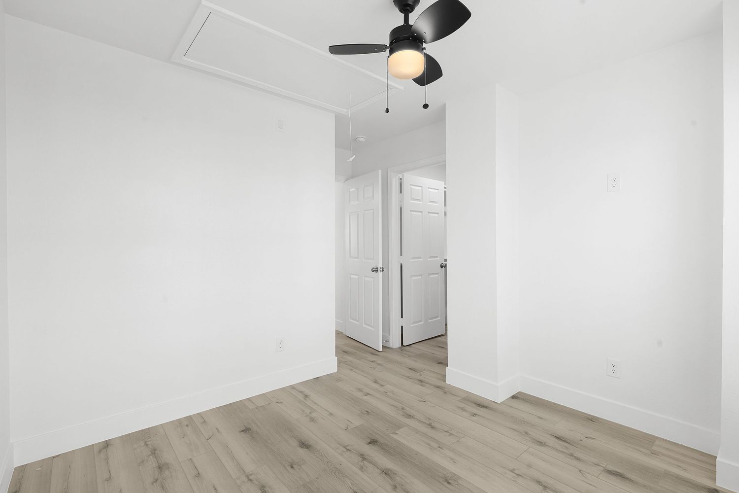 Empty white bedroom with light hardwood flooring and modern ceiling fan.