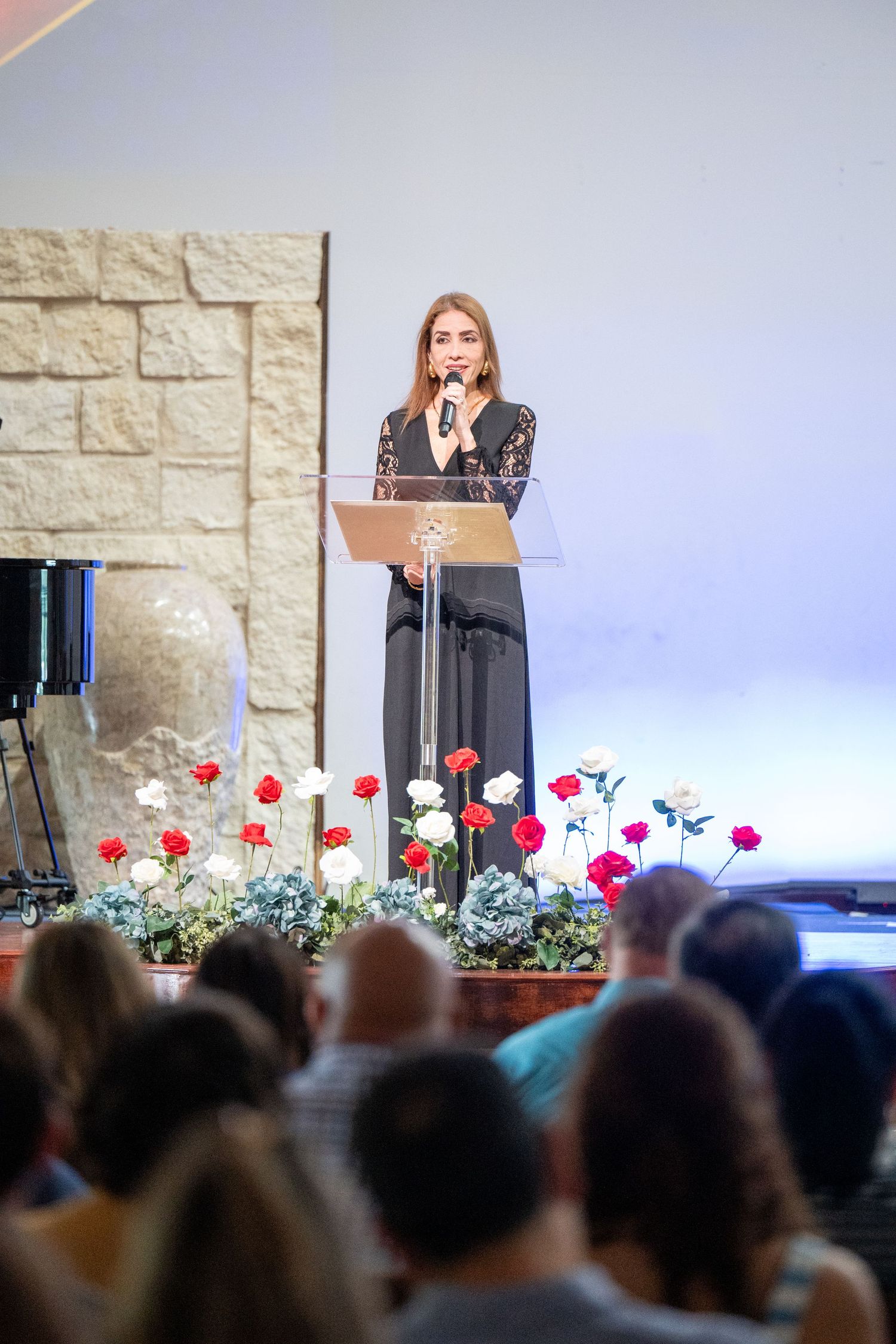 A speaker stands at a podium on stage decorated with colorful flowers while addressing an audience in an event venue.