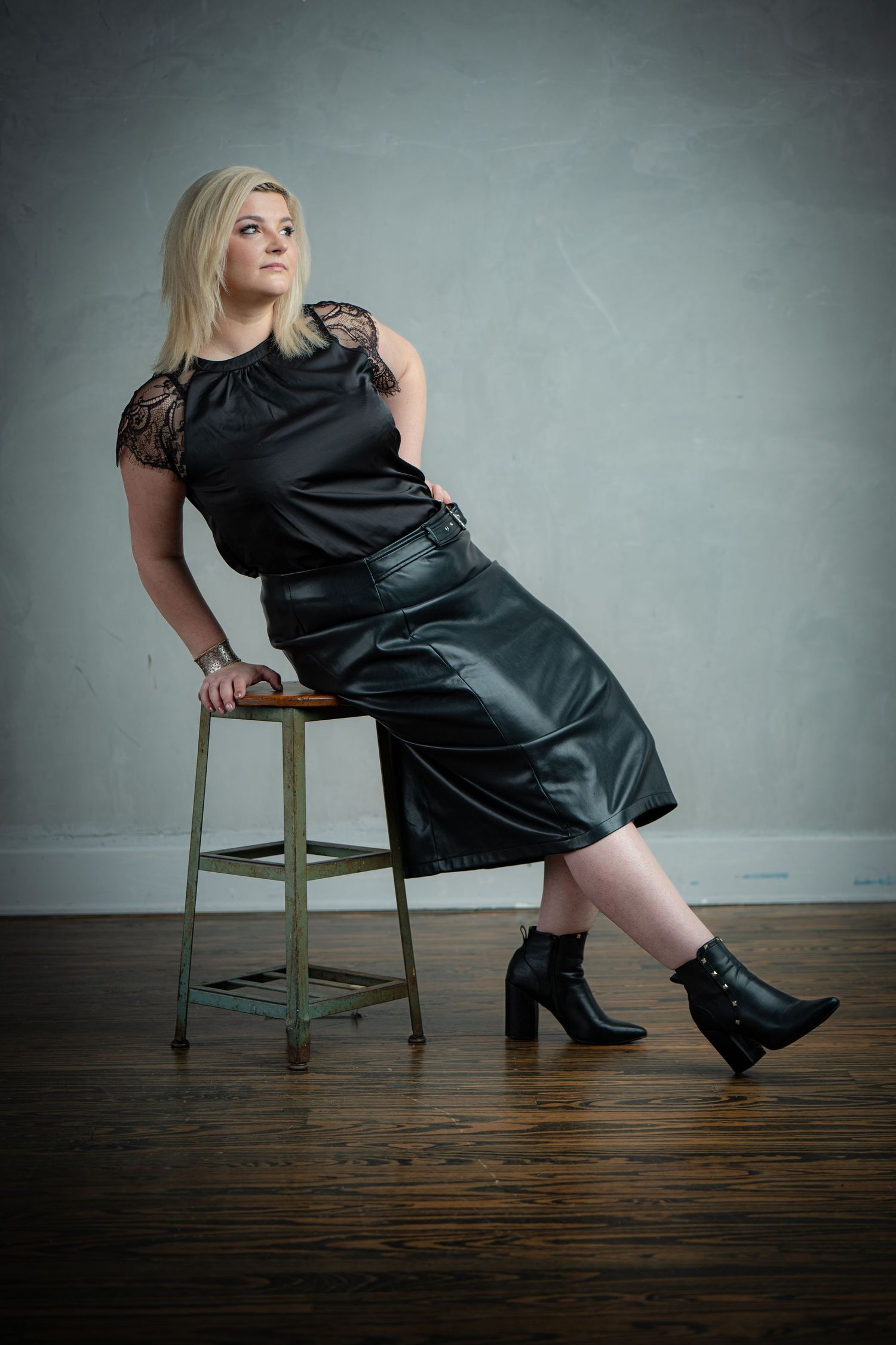 Woman in black dress and boots poses on wooden stool against grey wall in artistic photo series.