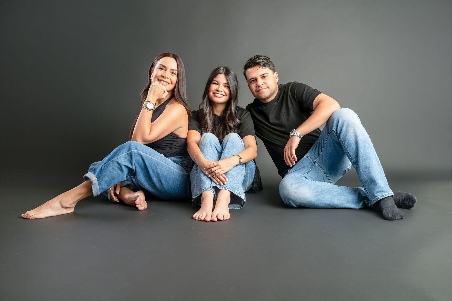 Three young adults sitting together on a studio floor, smiling, wearing casual jeans and tops against a gray background.