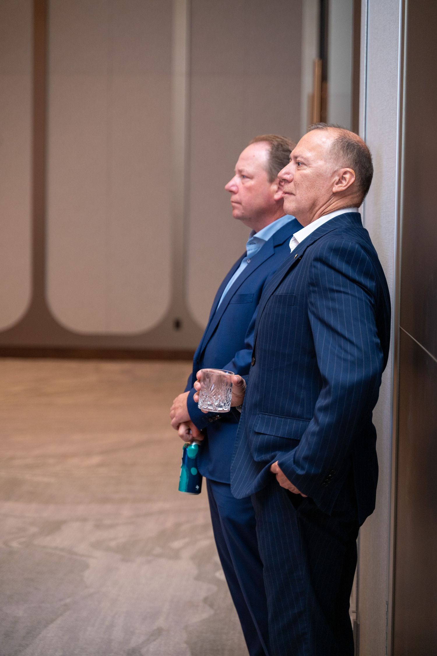 Two men in navy pinstripe suits stand side by side looking in the same direction inside a room with hardwood floors.