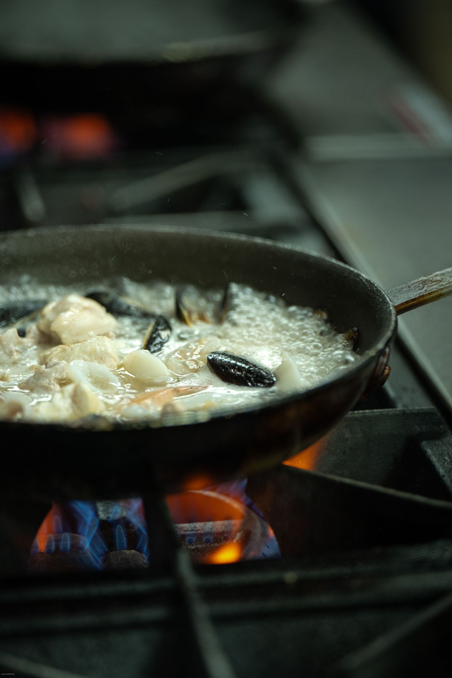 Cast iron pan cooking eggs over blue gas flame burners in professional kitchen.