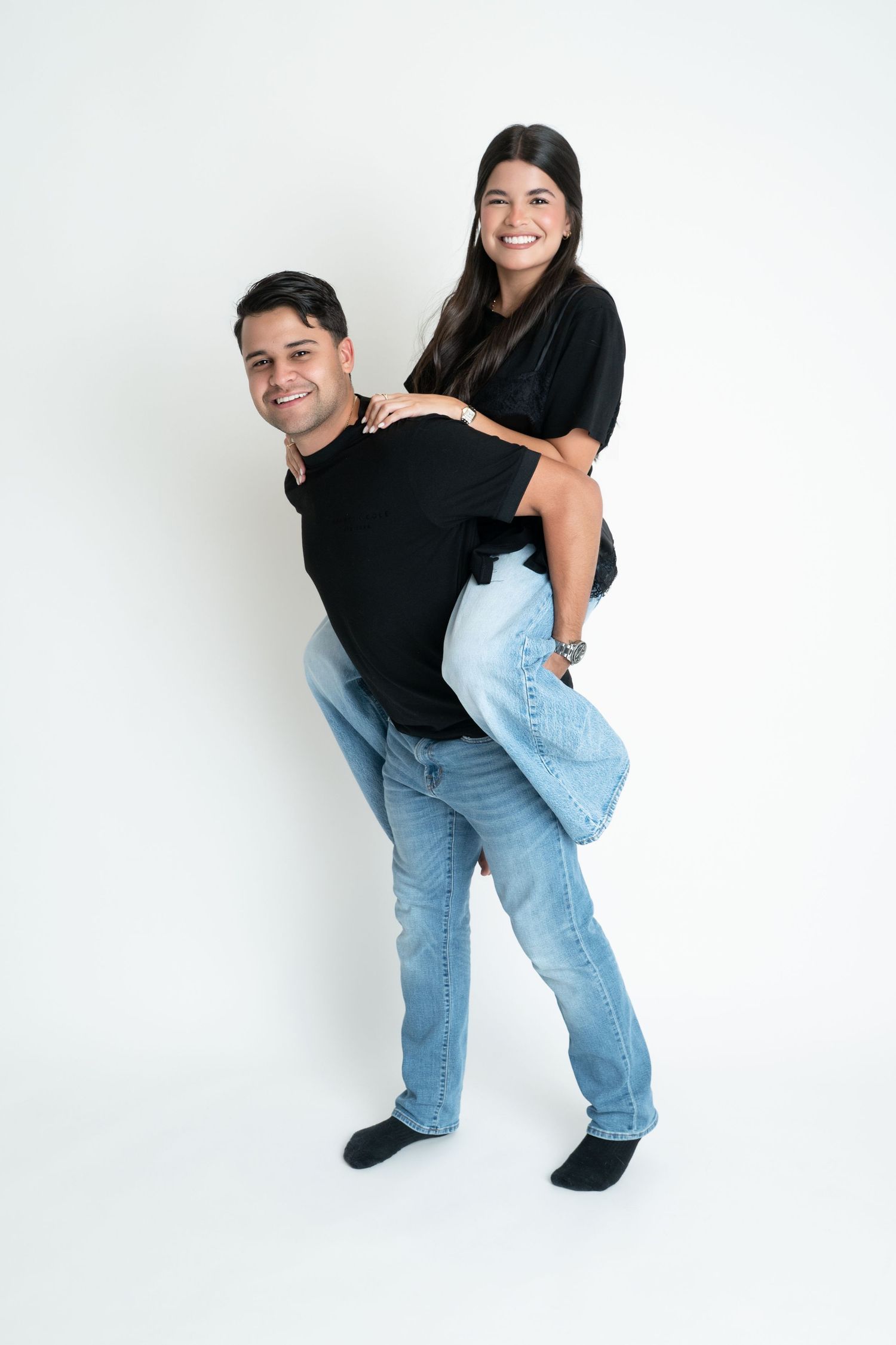 A smiling man gives a happy young woman a piggyback ride, both wearing black tops and jeans on white background.
