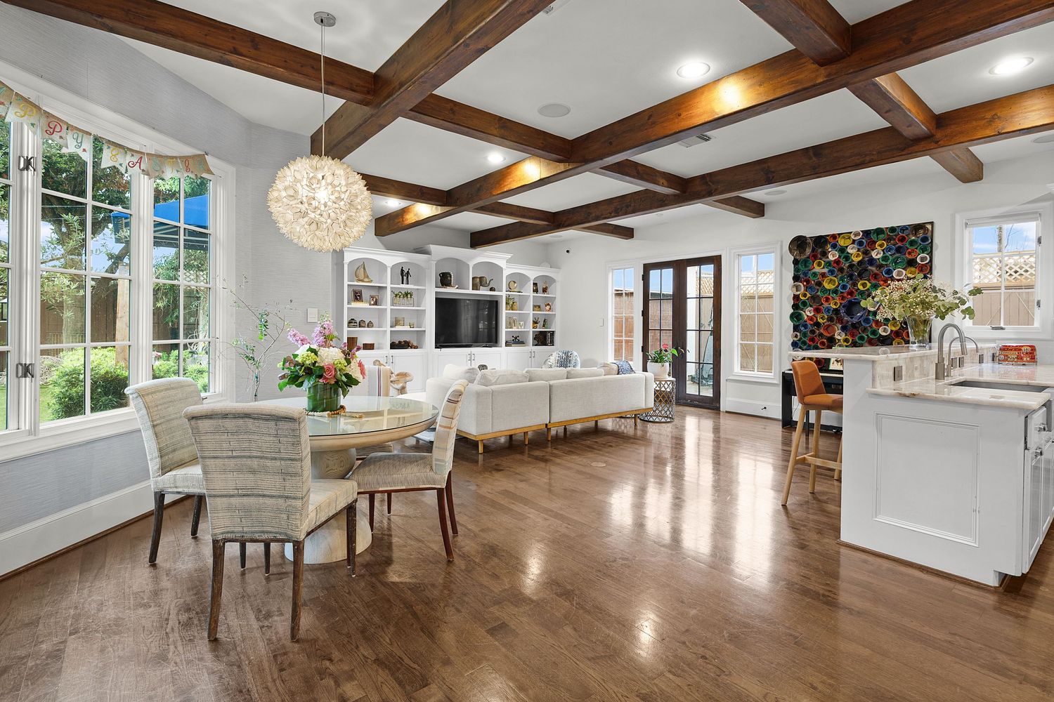Bright open-plan living and dining room with coffered wood beam ceiling, hardwood floors, built-in shelves, and French doors.