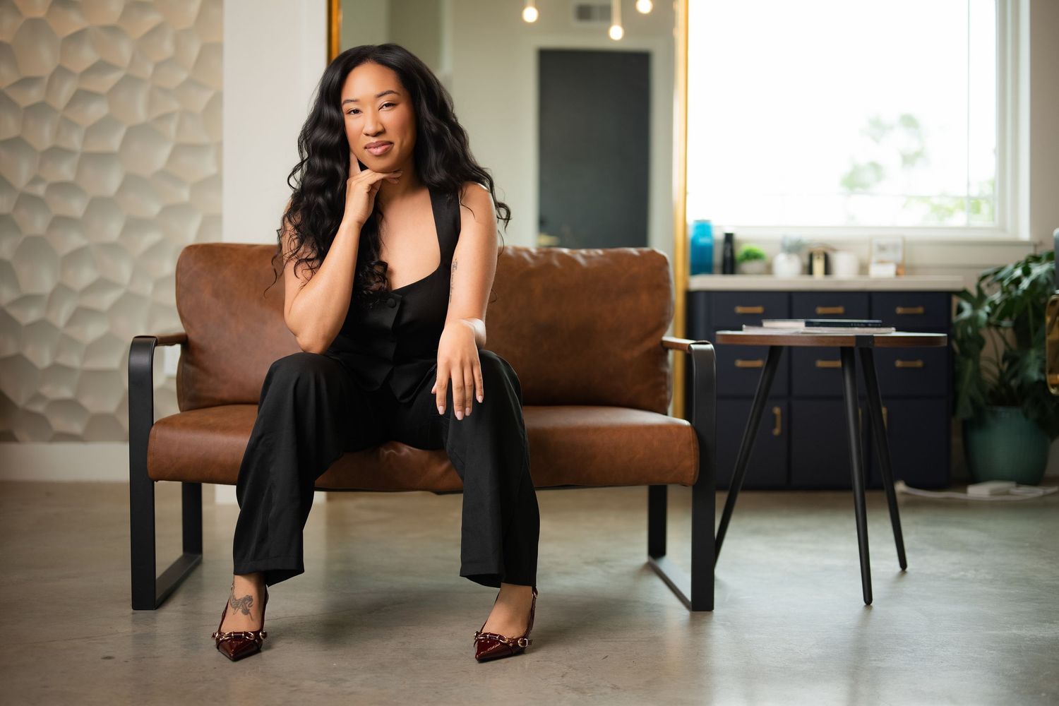 Person in black outfit sitting on brown leather couch in modern salon setting.