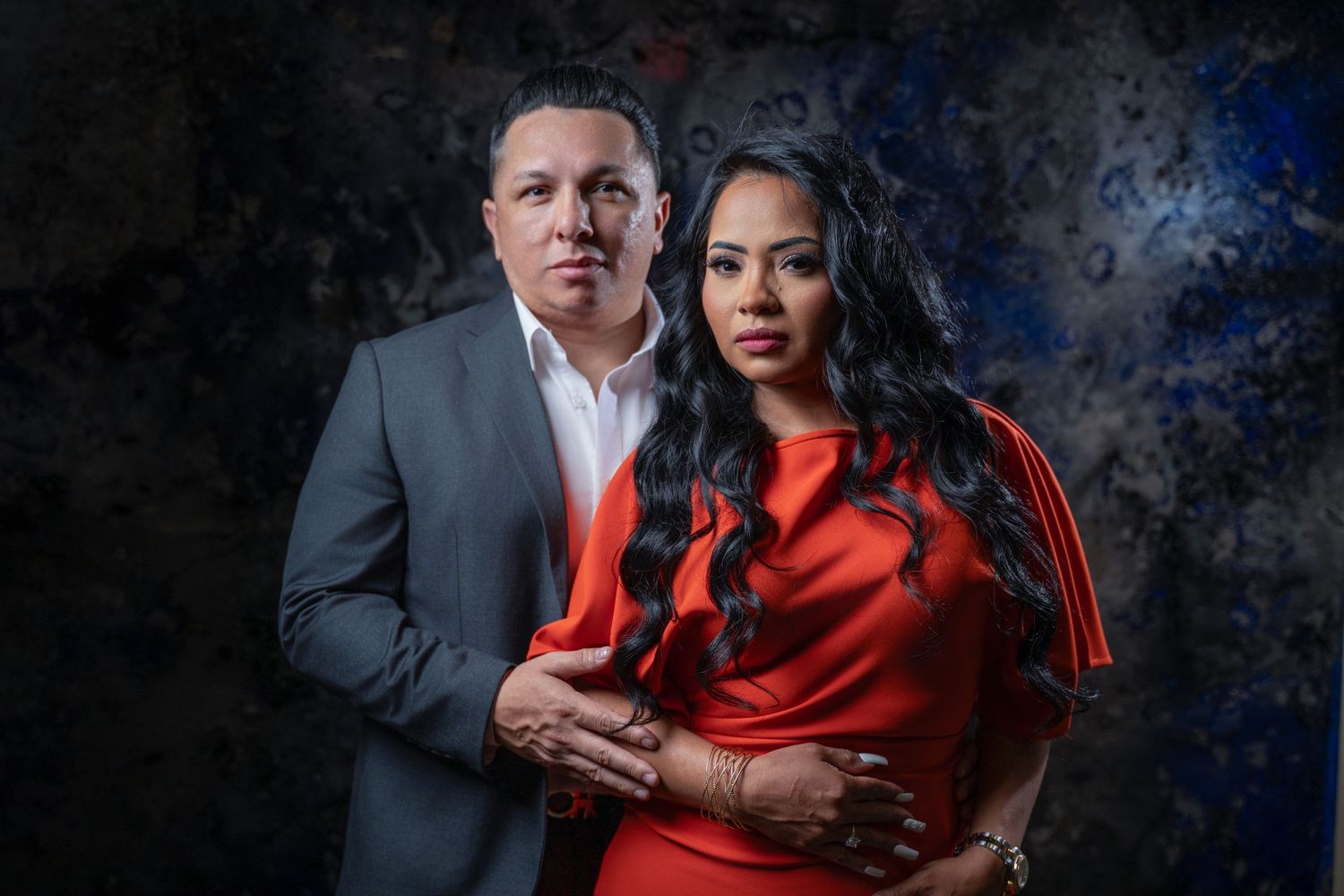 A romantic couple poses together against a dark backdrop in a professional studio portrait with red formal attire.