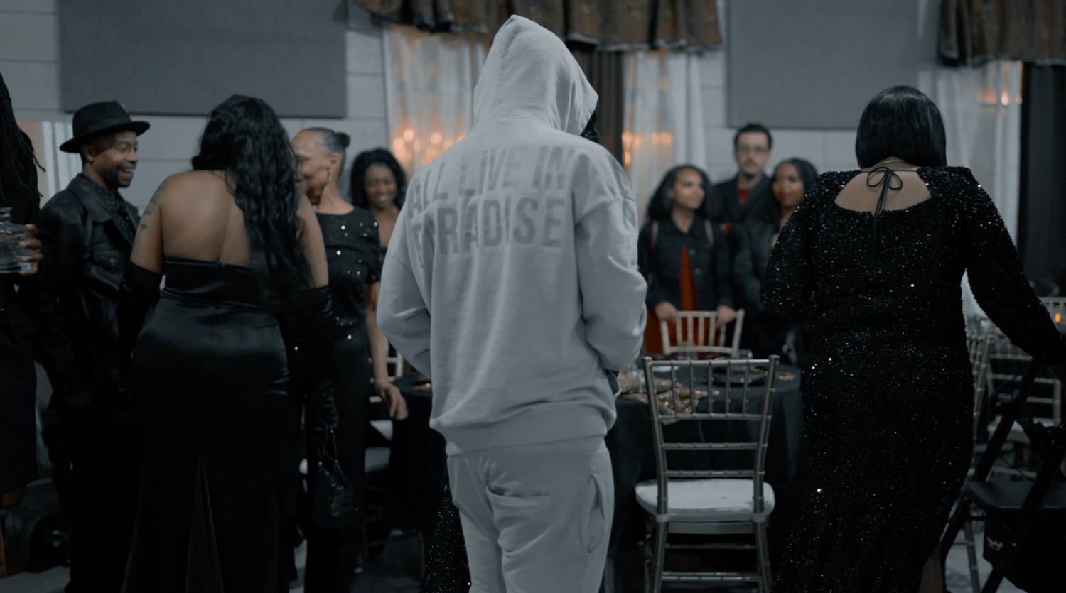 A group of people gathered inside a dimly lit venue with someone wearing a white hoodie in the center.