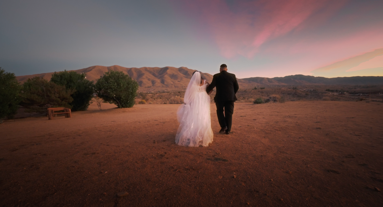 A couple in wedding attire walking into the desert sunset with mountains and pink sky in the background.
