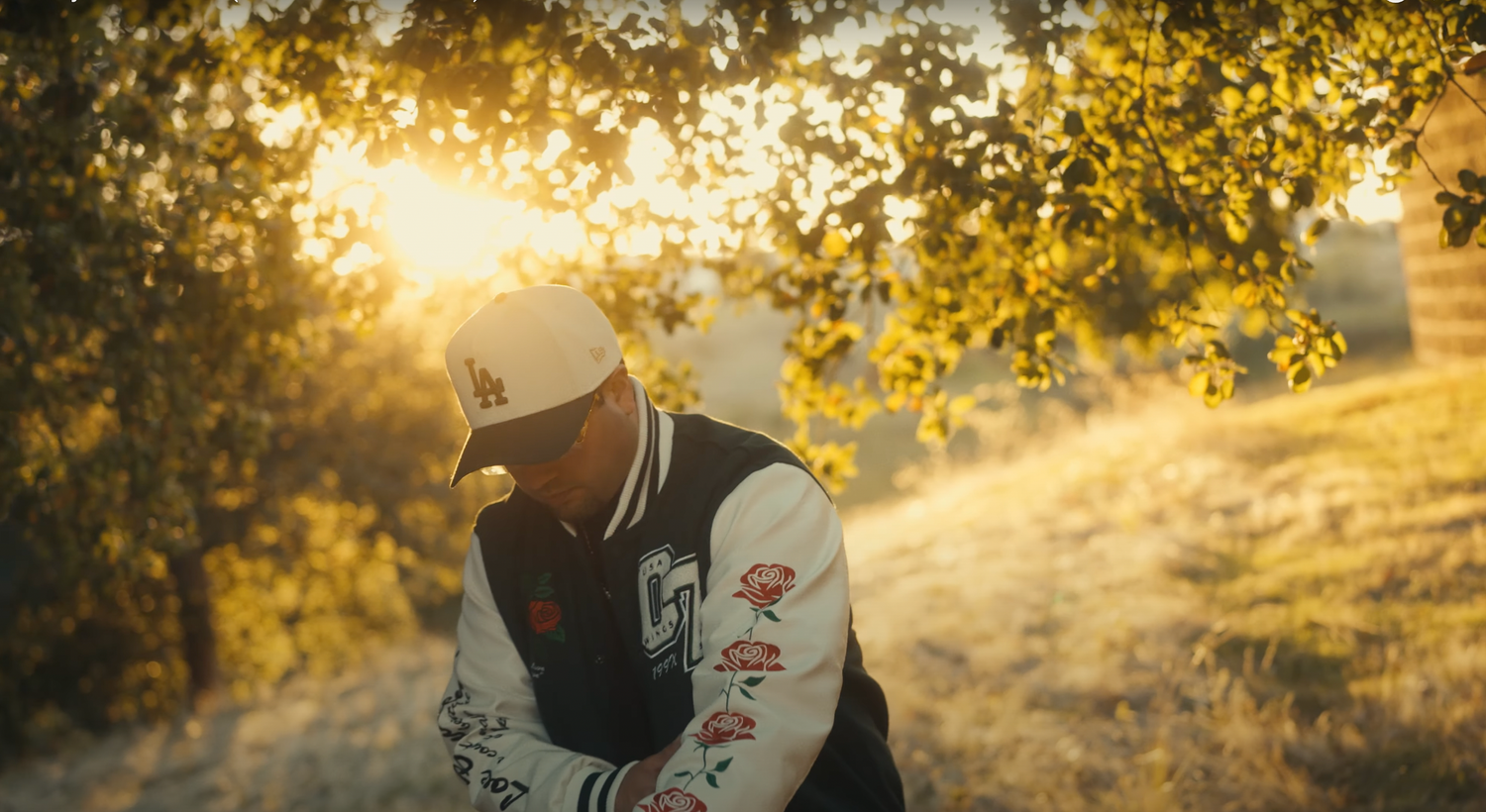 A person in a varsity jacket sits outdoors during golden hour sunset with sunlight streaming through autumn leaves.