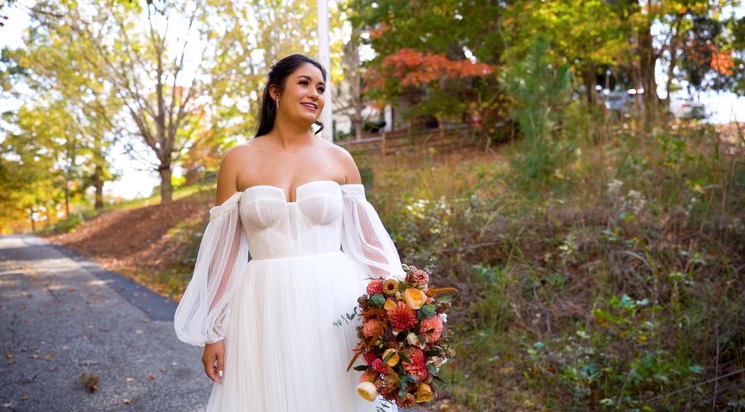 A bride in a flowing white gown holds an autumn bouquet while walking along a sunlit outdoor path.
