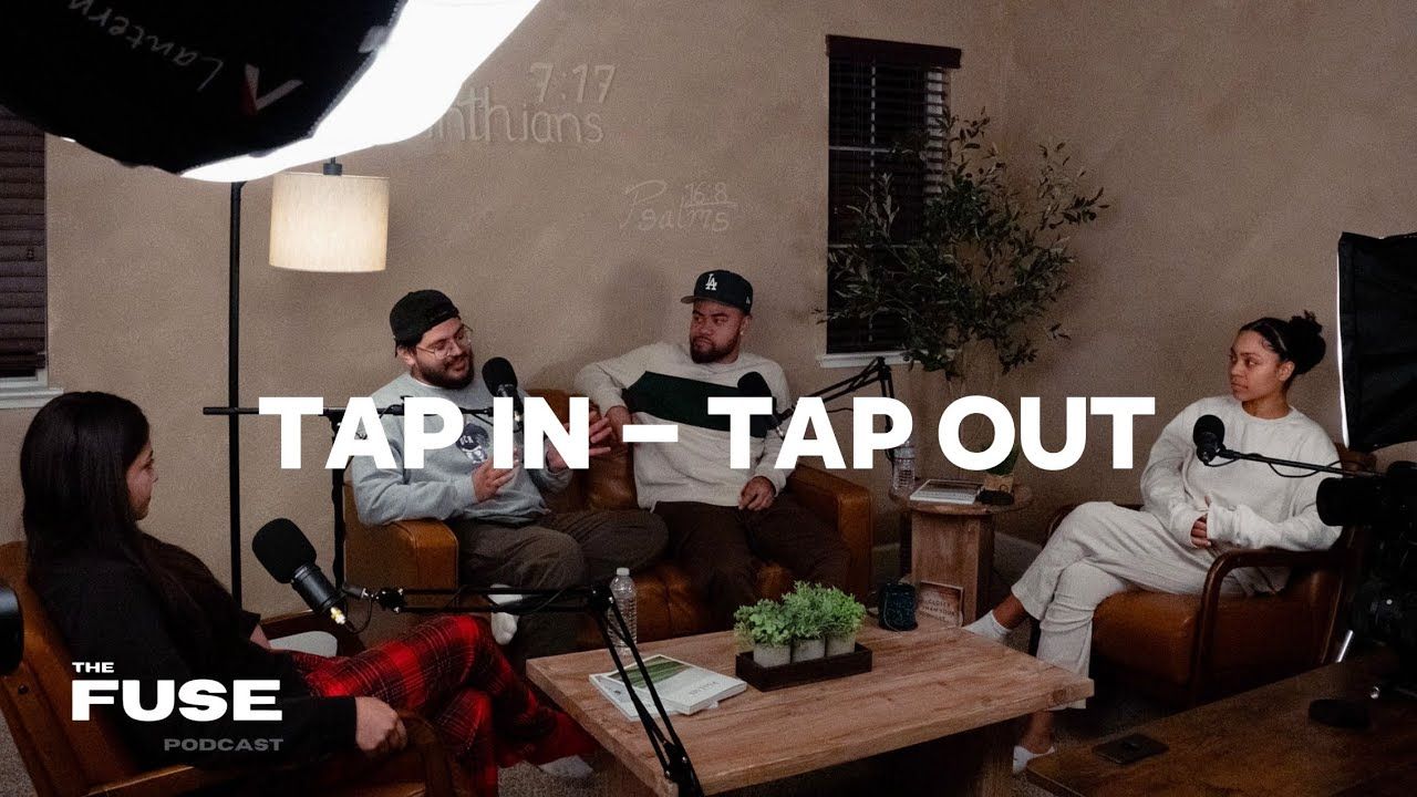 Four people sit at a wooden outdoor table having a casual conversation under a tap in tap out text overlay.
