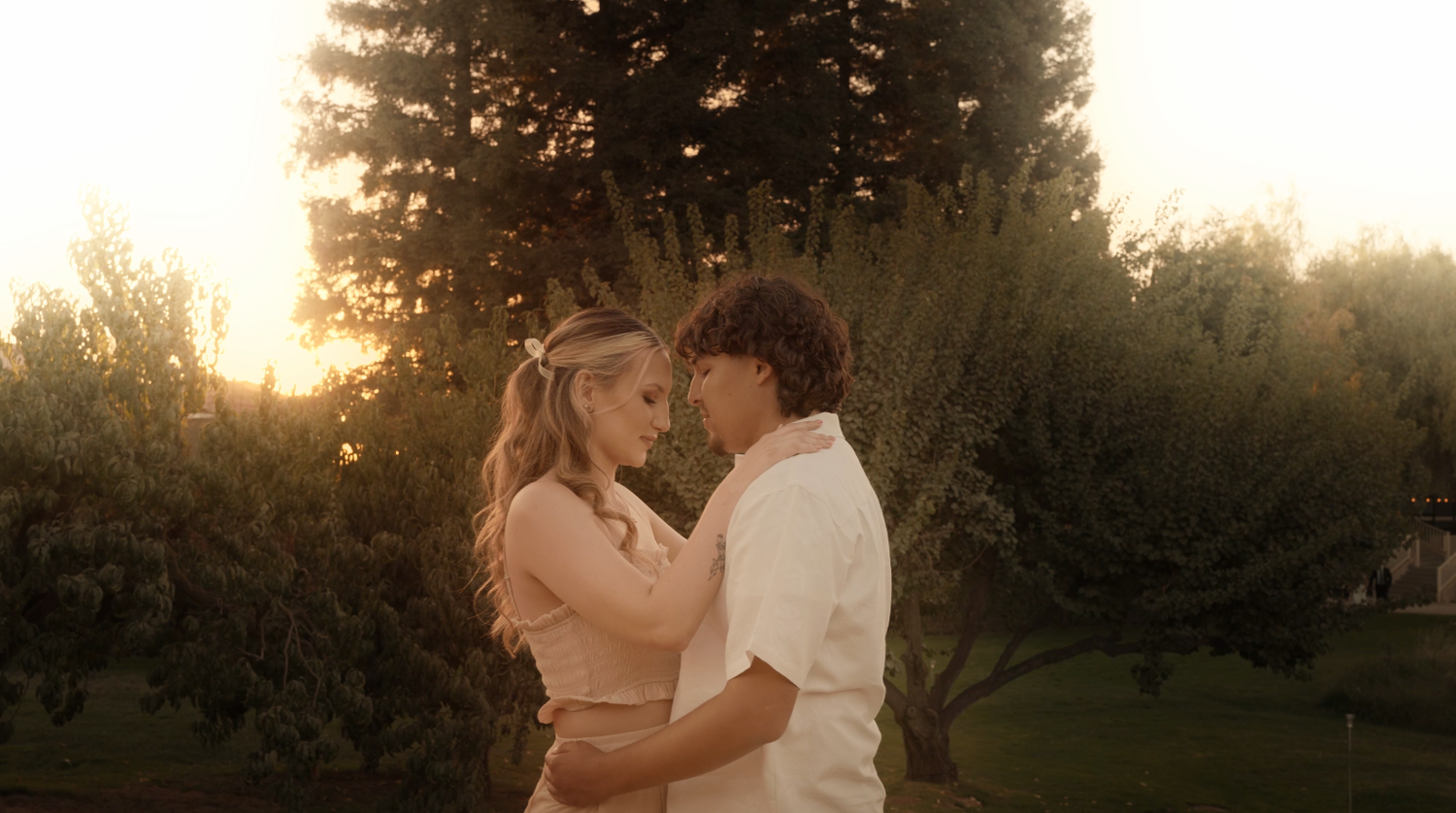 A romantic couple embraces in a garden at golden hour with sunlit trees creating a dreamy backdrop.