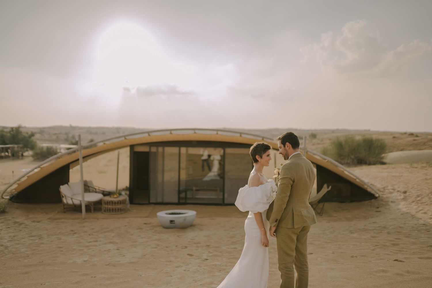 A couple in wedding attire stands in front of a curved architectural structure in the desert landscape.