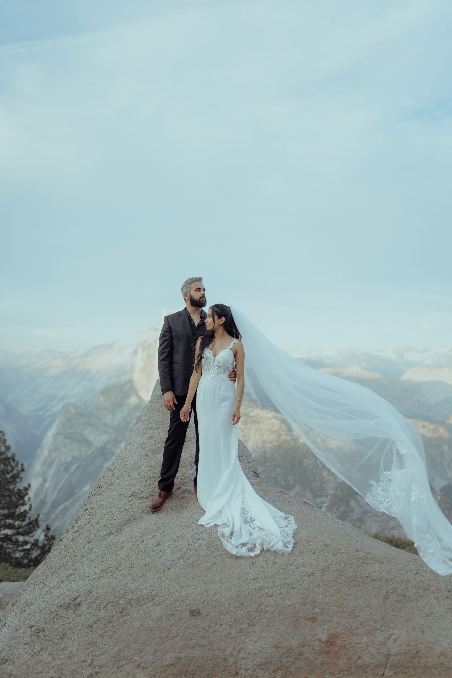 Wedding couple standing on mountain peak with flowing veil against dramatic landscape.