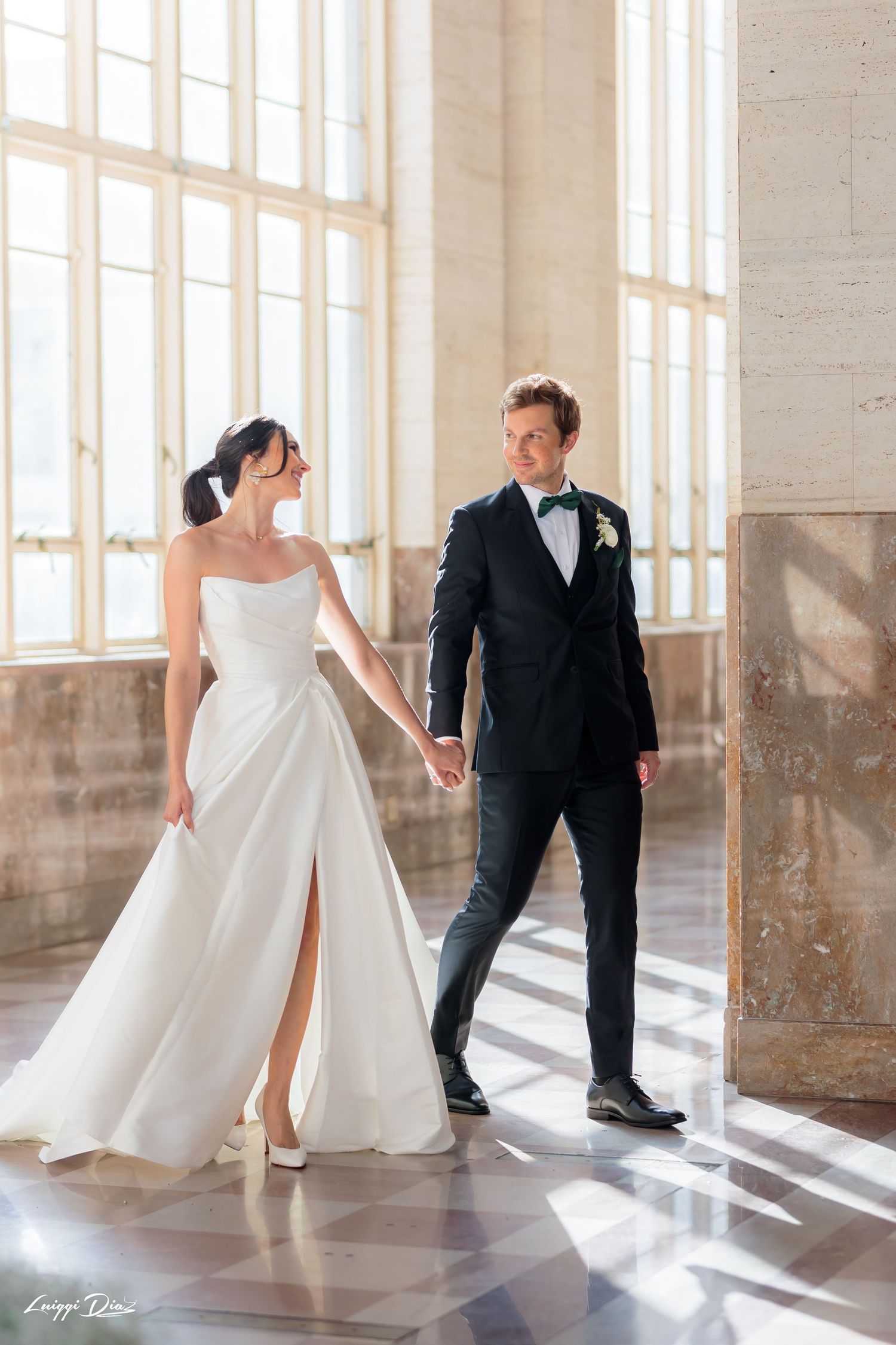 Elegant couple in wedding attire holding hands in a bright room with tall windows.
