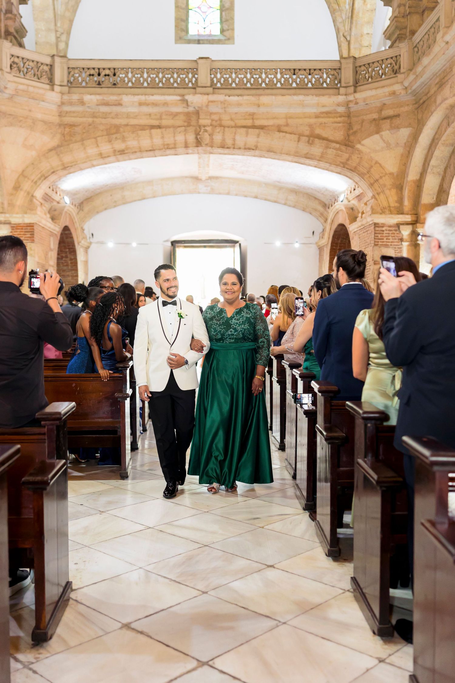 Wedding guests photograph couple walking down church aisle with ornate stone architecture.