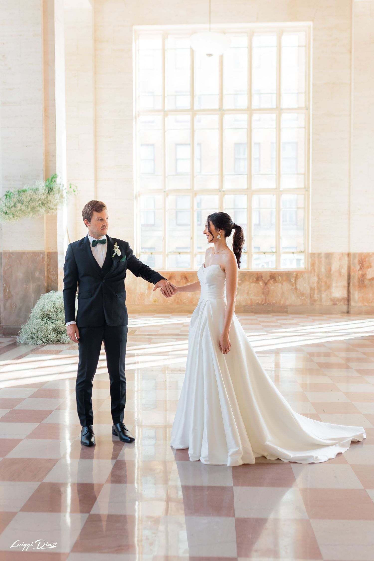 Couple in wedding attire holding hands in an elegant venue with tall windows.