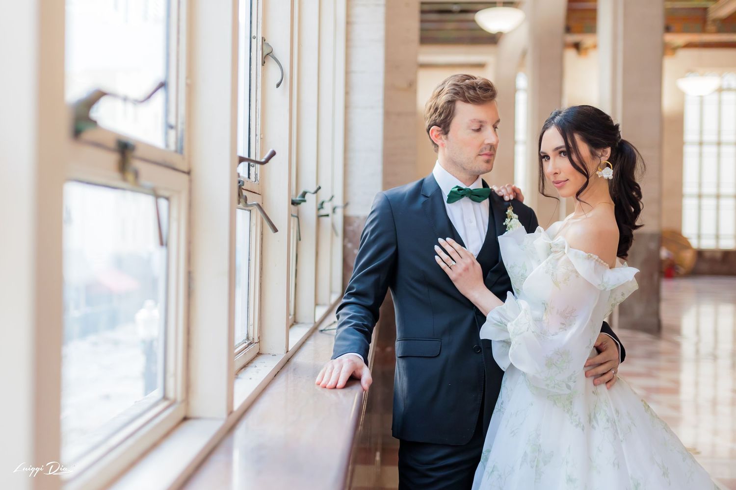 Romantic couple pose by large windows in wedding attire.
