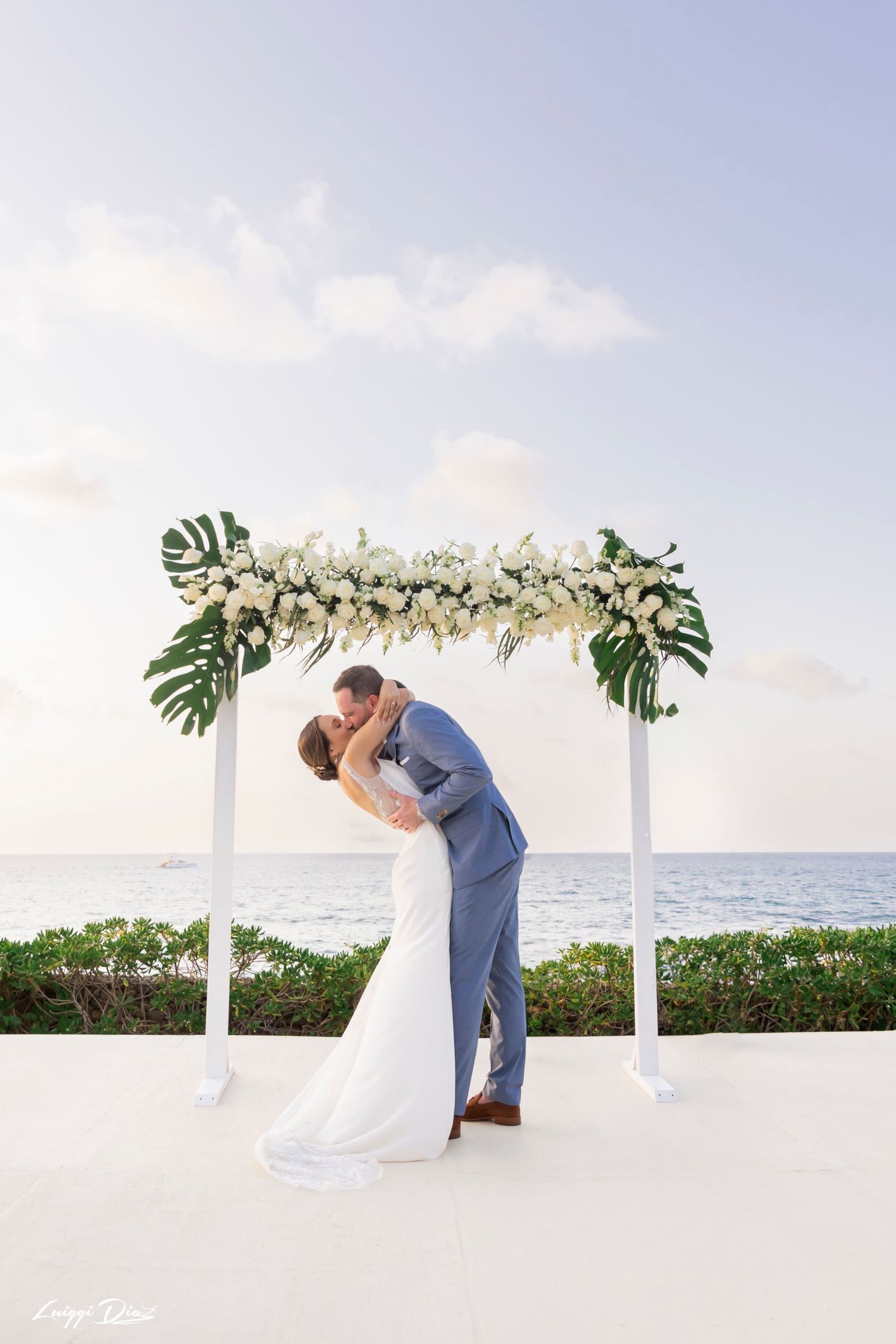 Romantic beach wedding ceremony under a white arch decorated with tropical leaves and flowers.