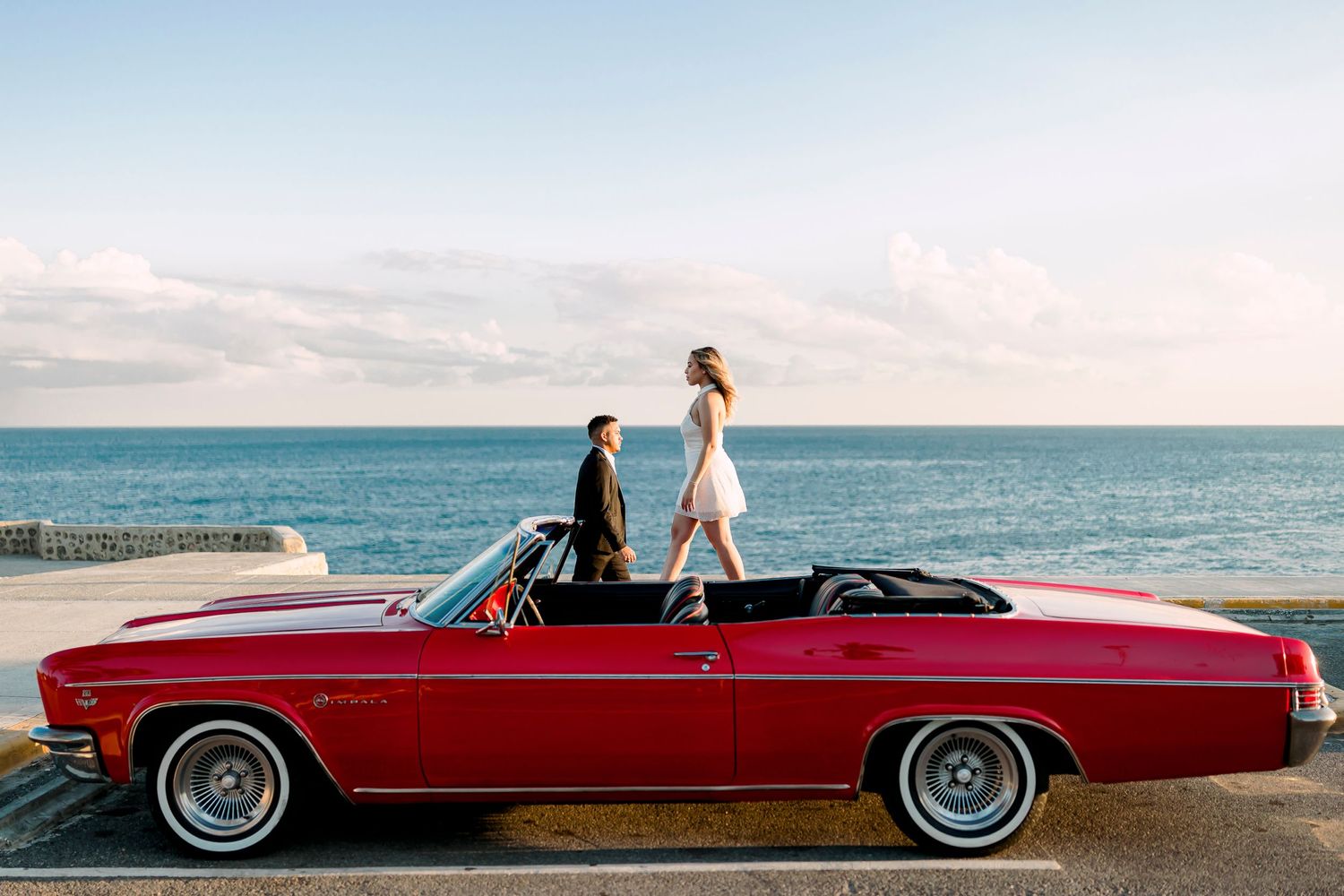 Red classic convertible parked along oceanfront with couple posing on car.
