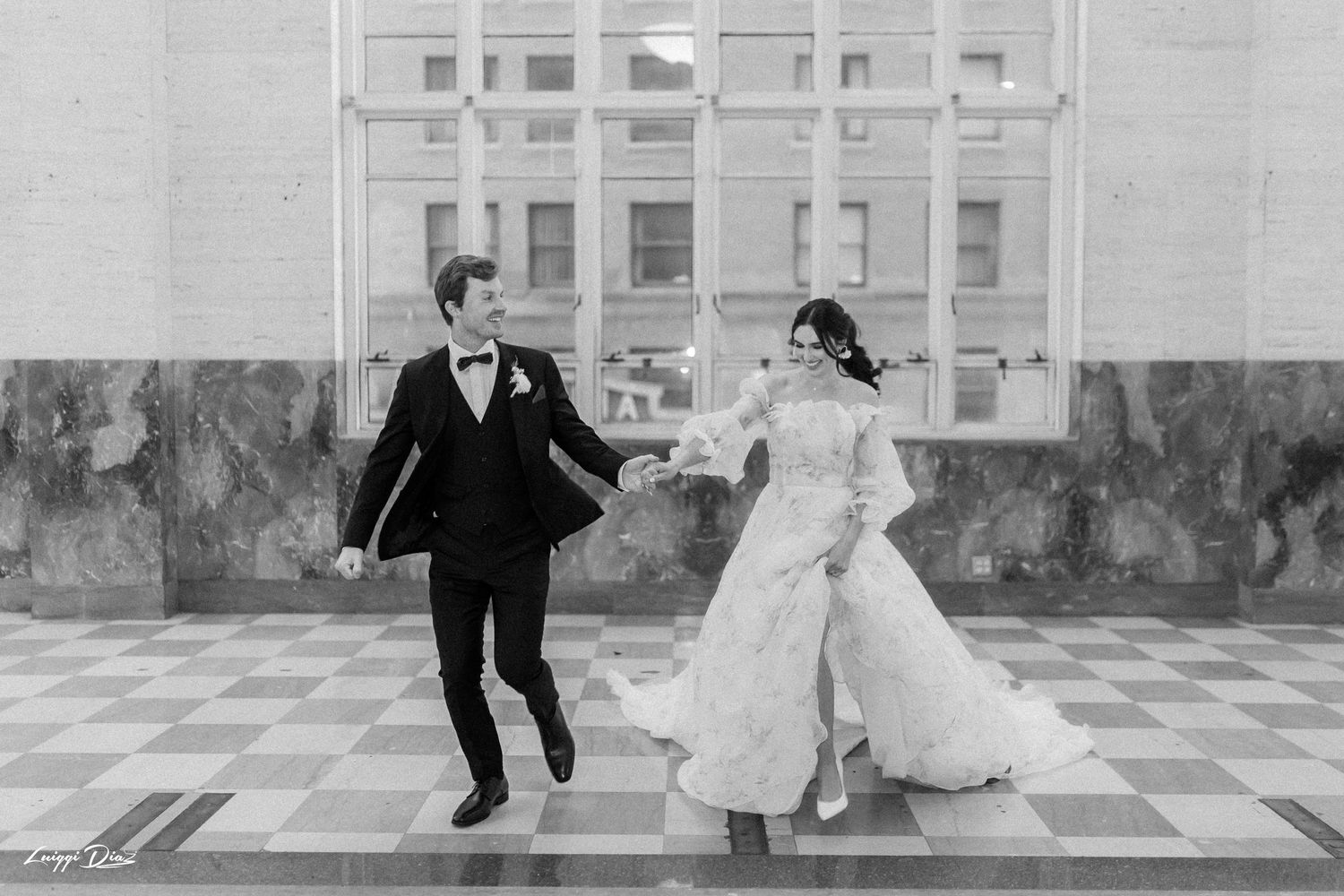 A couple in elegant wedding attire run joyfully together across a patterned outdoor terrace in black and white.