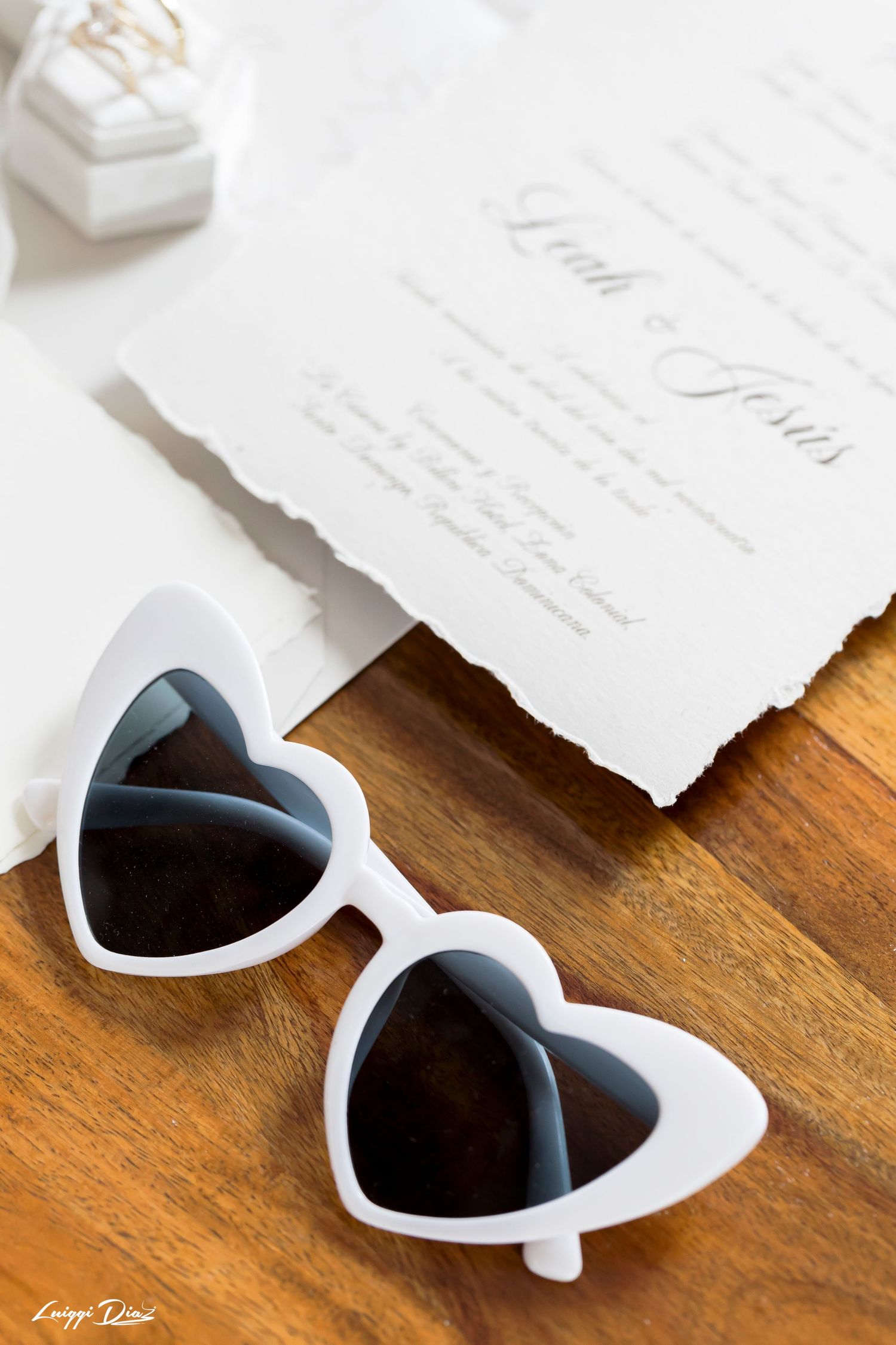 White heart-shaped sunglasses next to elegant wedding stationery on wooden surface.