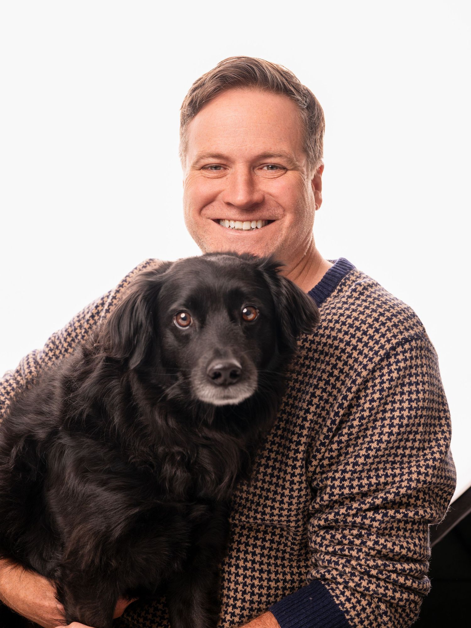 Smiling man in houndstooth sweater holding a fluffy black dog against a white background.