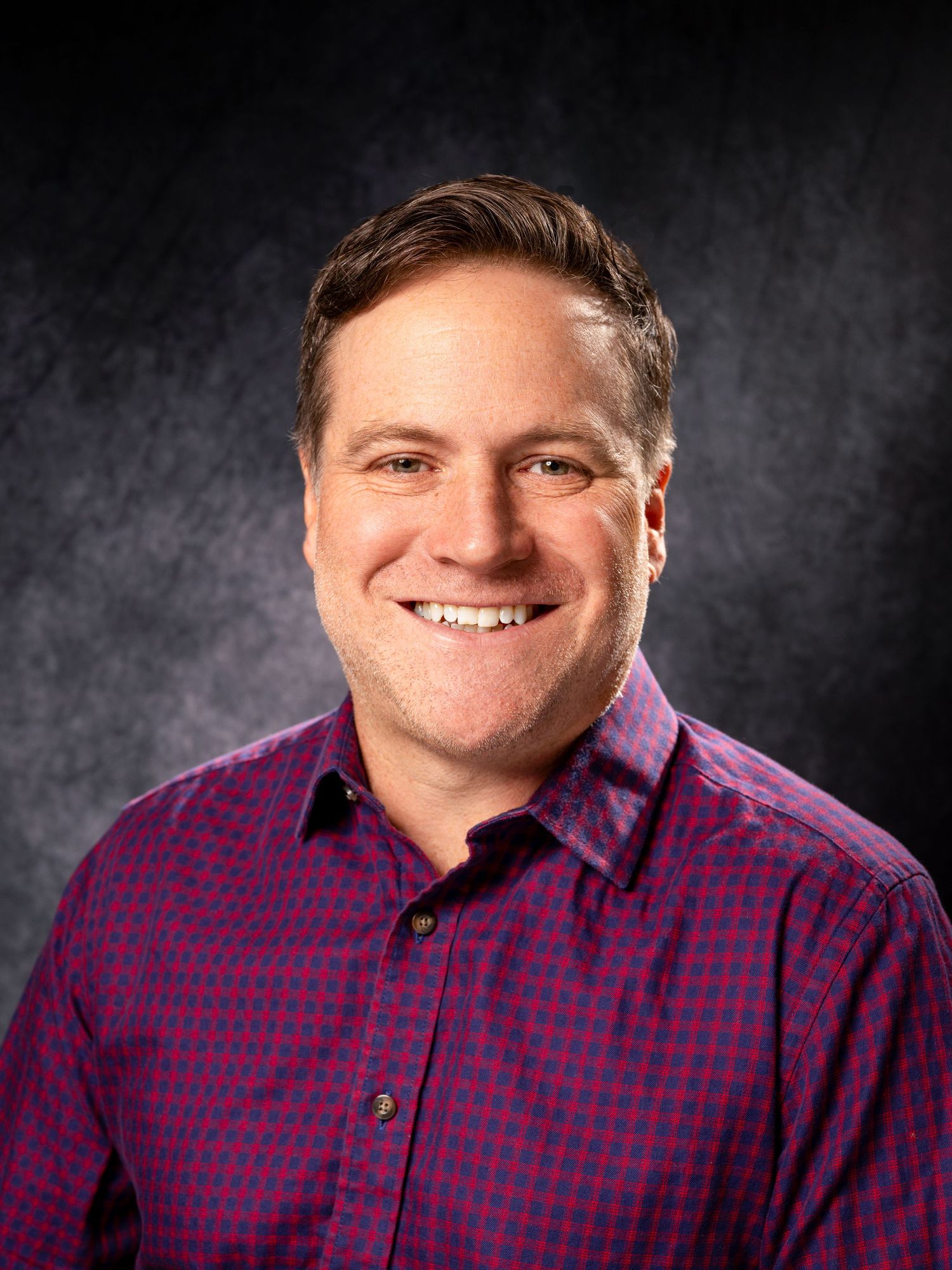 Professional headshot portrait against dark background wearing purple and black checkered button-down shirt.