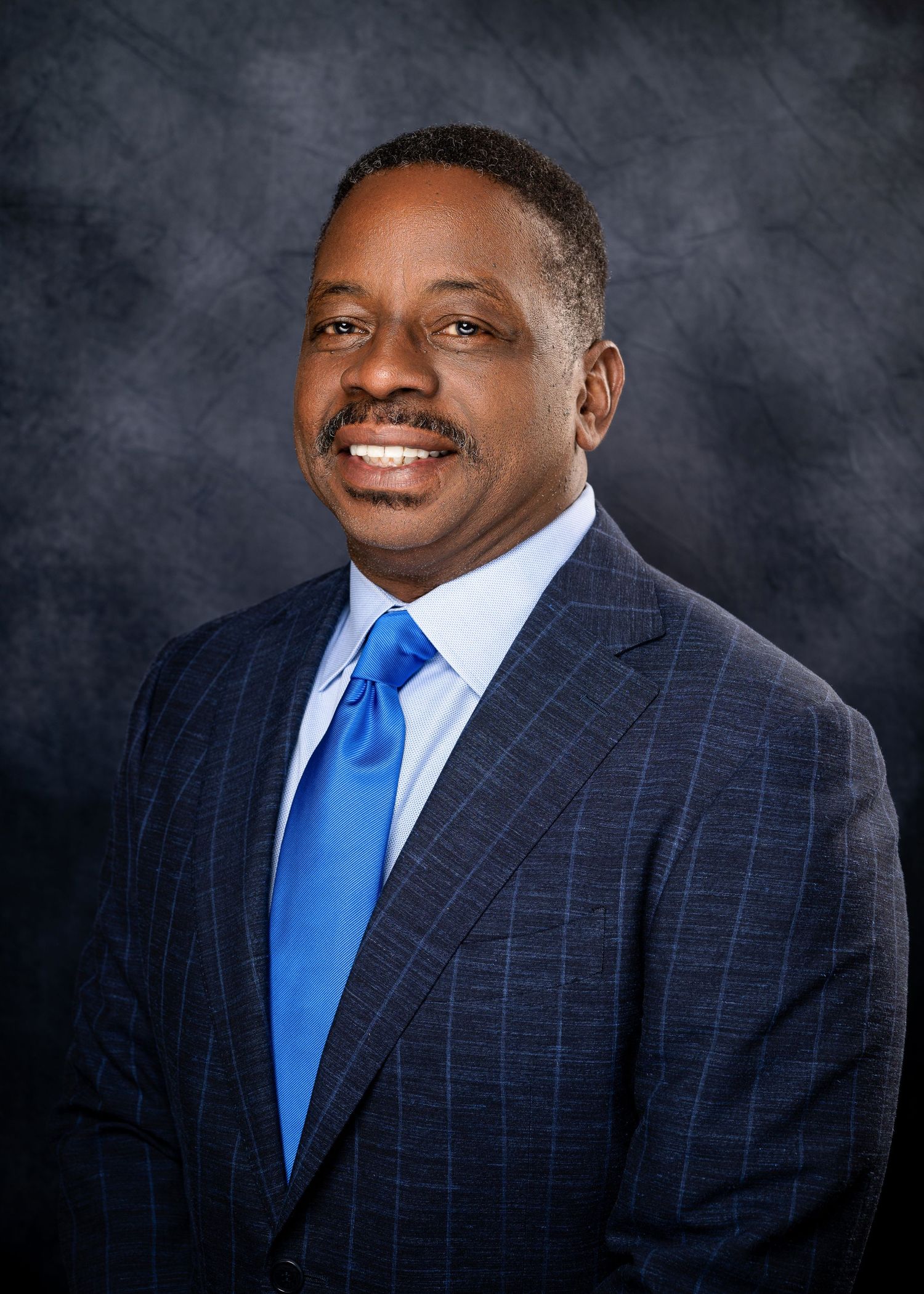 Professional portrait of someone in a navy windowpane suit and bright blue tie against a dark background.