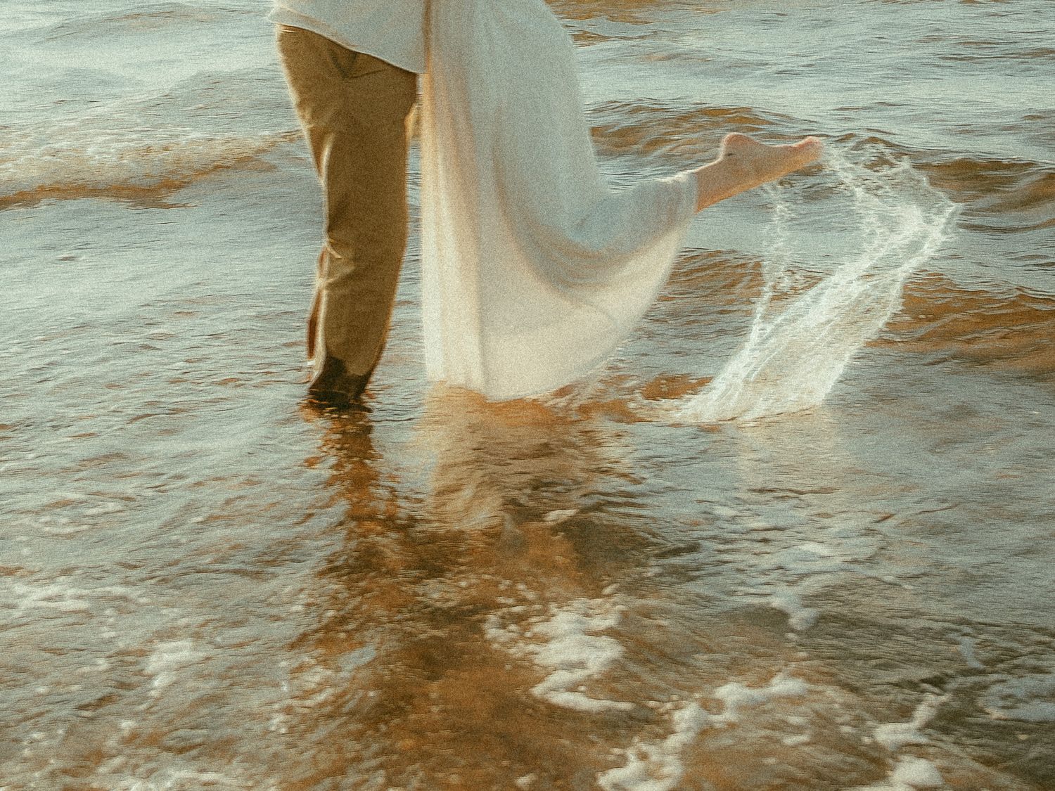 Couple walking in ocean waves, bride in flowing white dress with tan pants groom, romantic beach wedding photo.