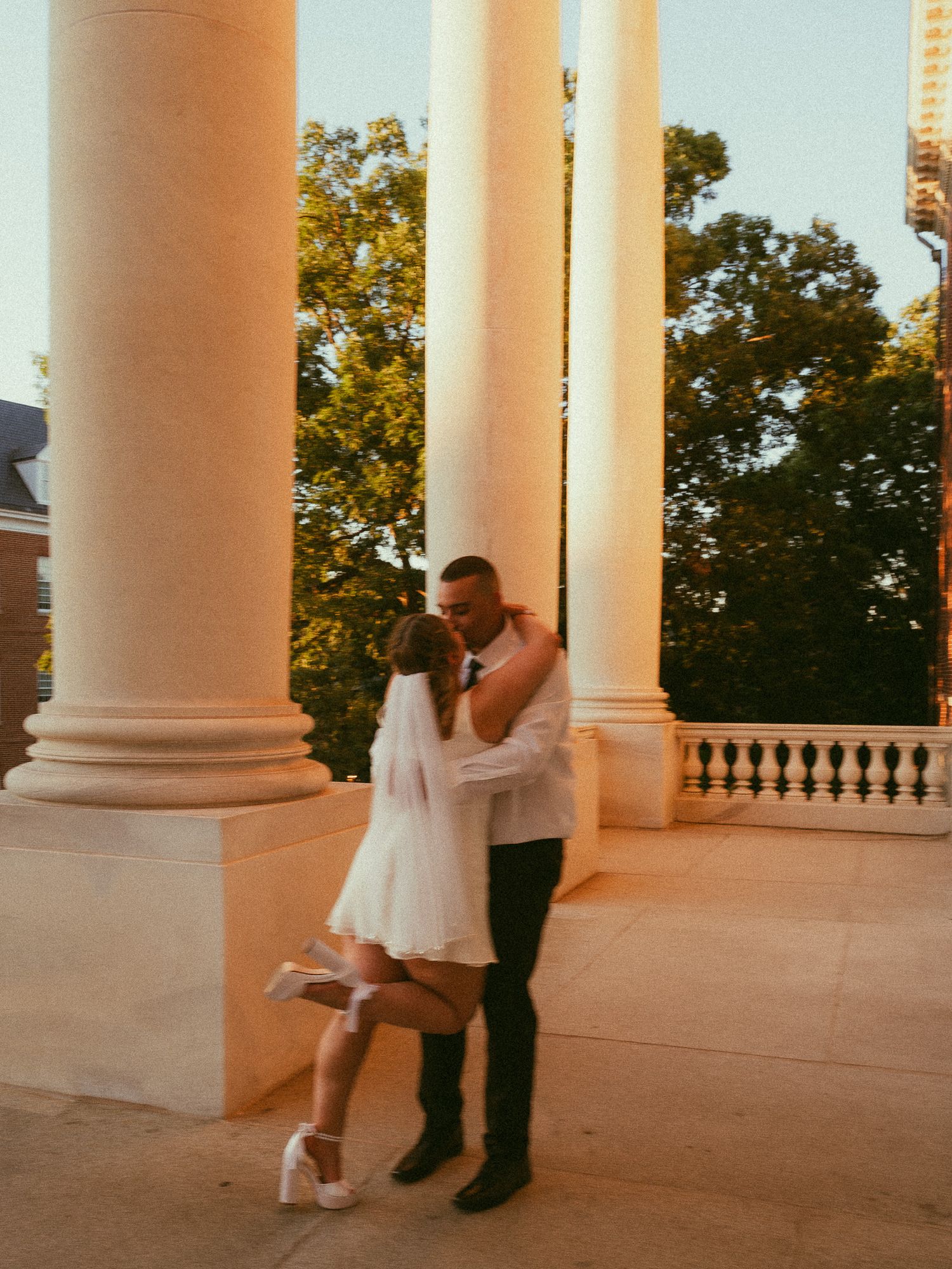 Couple sharing a romantic kiss in wedding attire among white columns at golden hour.