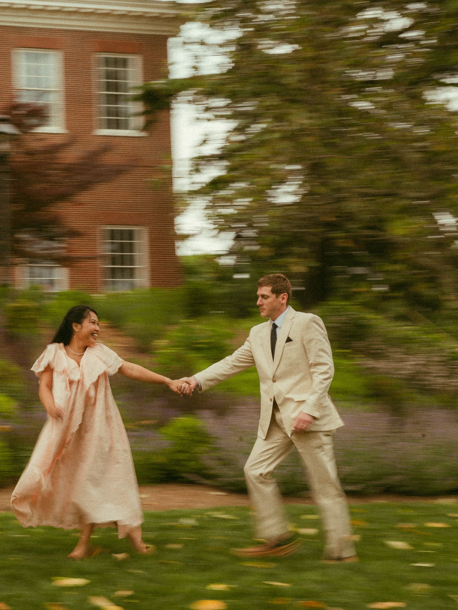 Couple holding hands dancing on green lawn near brick building, woman in pink dress, man in cream suit.