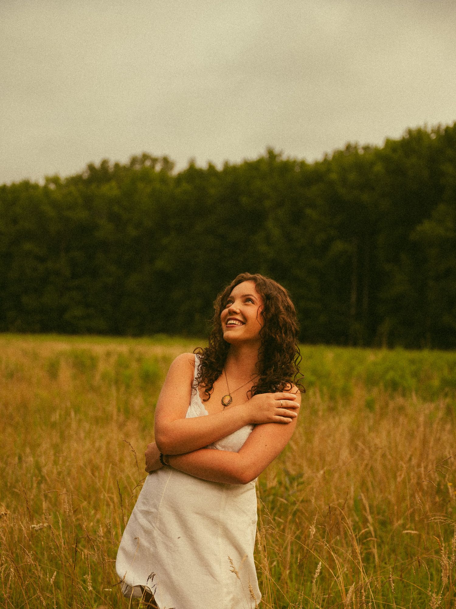 A smiling woman in a white dress stands in a golden meadow with arms crossed, surrounded by lush green trees.