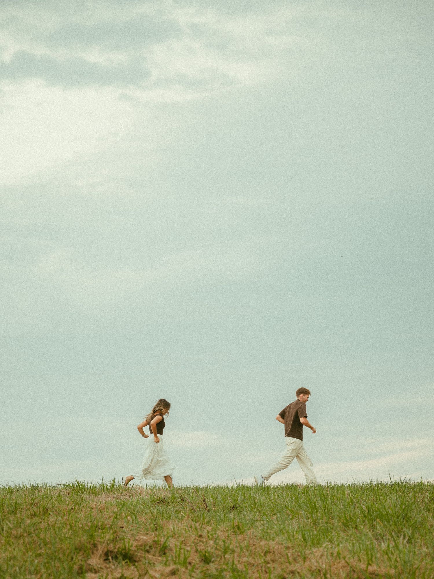 Two people walking across a grassy hill under a cloudy sky, one in a white skirt and one in light pants.