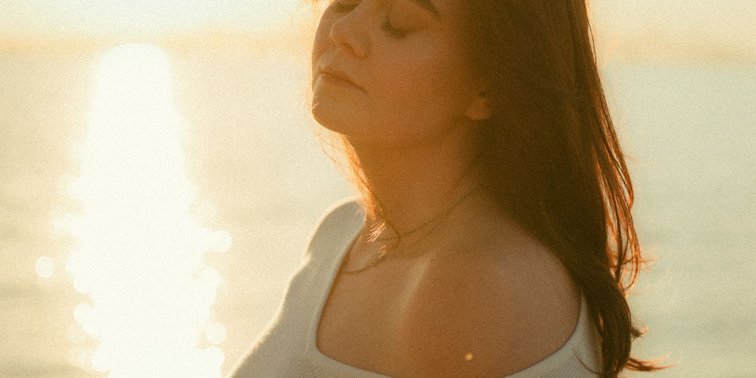 Woman with eyes closed basking in golden sunset light by the ocean, wearing a white top and delicate necklace.
