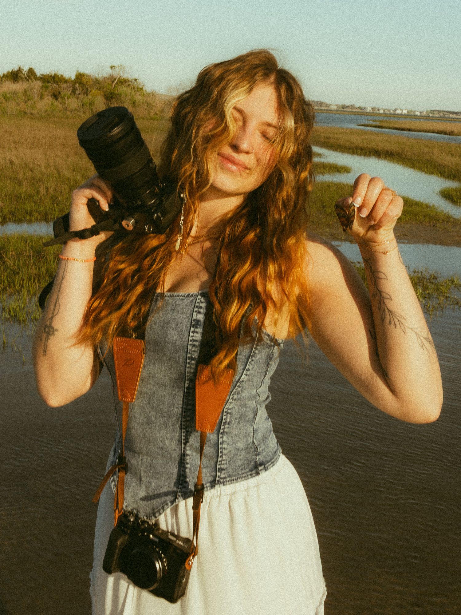 Young woman with wavy auburn hair holds a camera outdoors near a marsh, wearing a denim corset and white skirt.