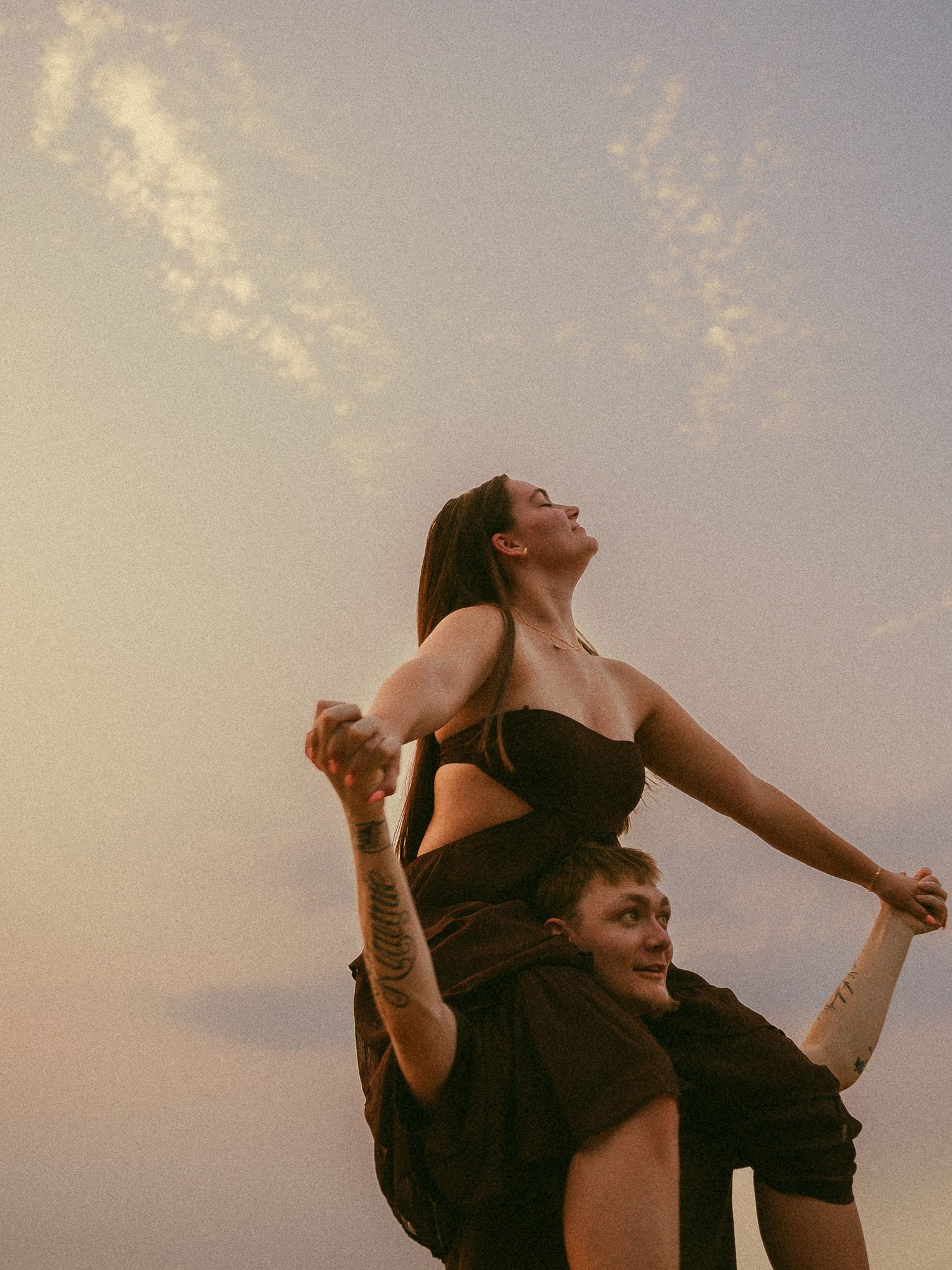 A woman in a black dress sits on a man's shoulders against a pastel sunset sky, arms outstretched joyfully.