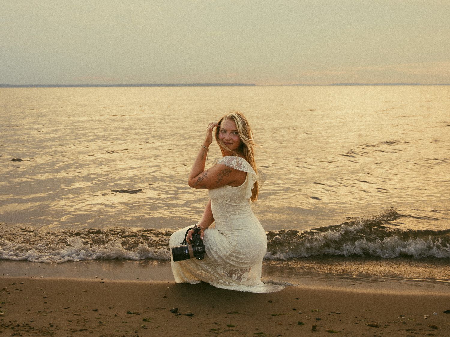 Woman in white lace dress posing on sandy beach at sunset with calm ocean waves behind her.