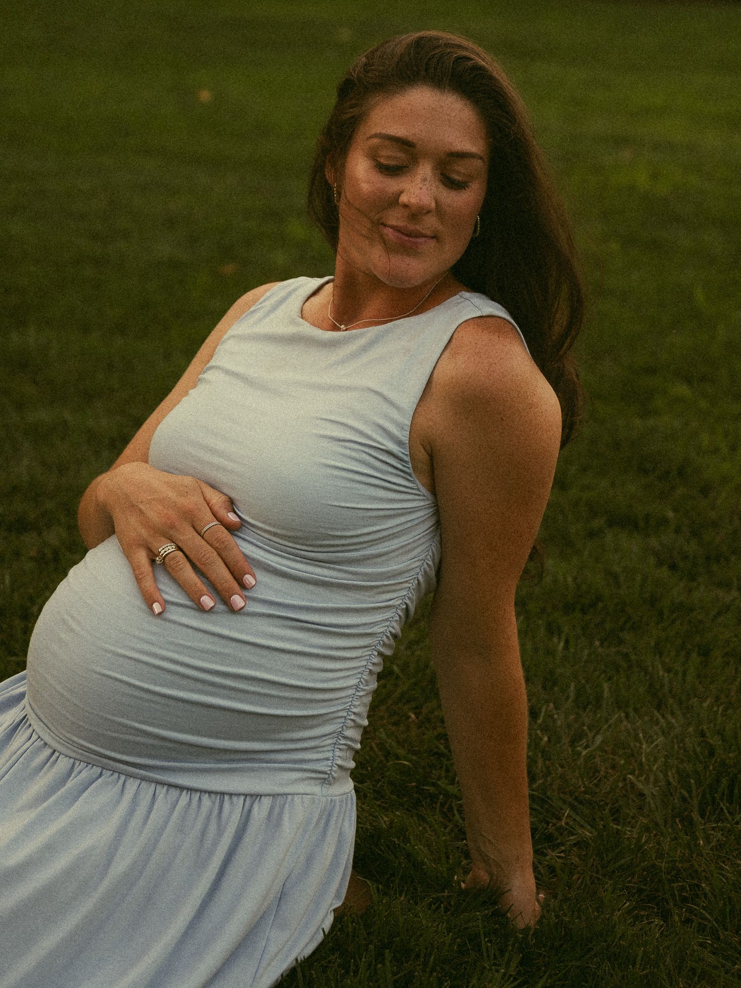 Pregnant woman in light blue ruched dress sitting on grass, cradling her baby bump outdoors.