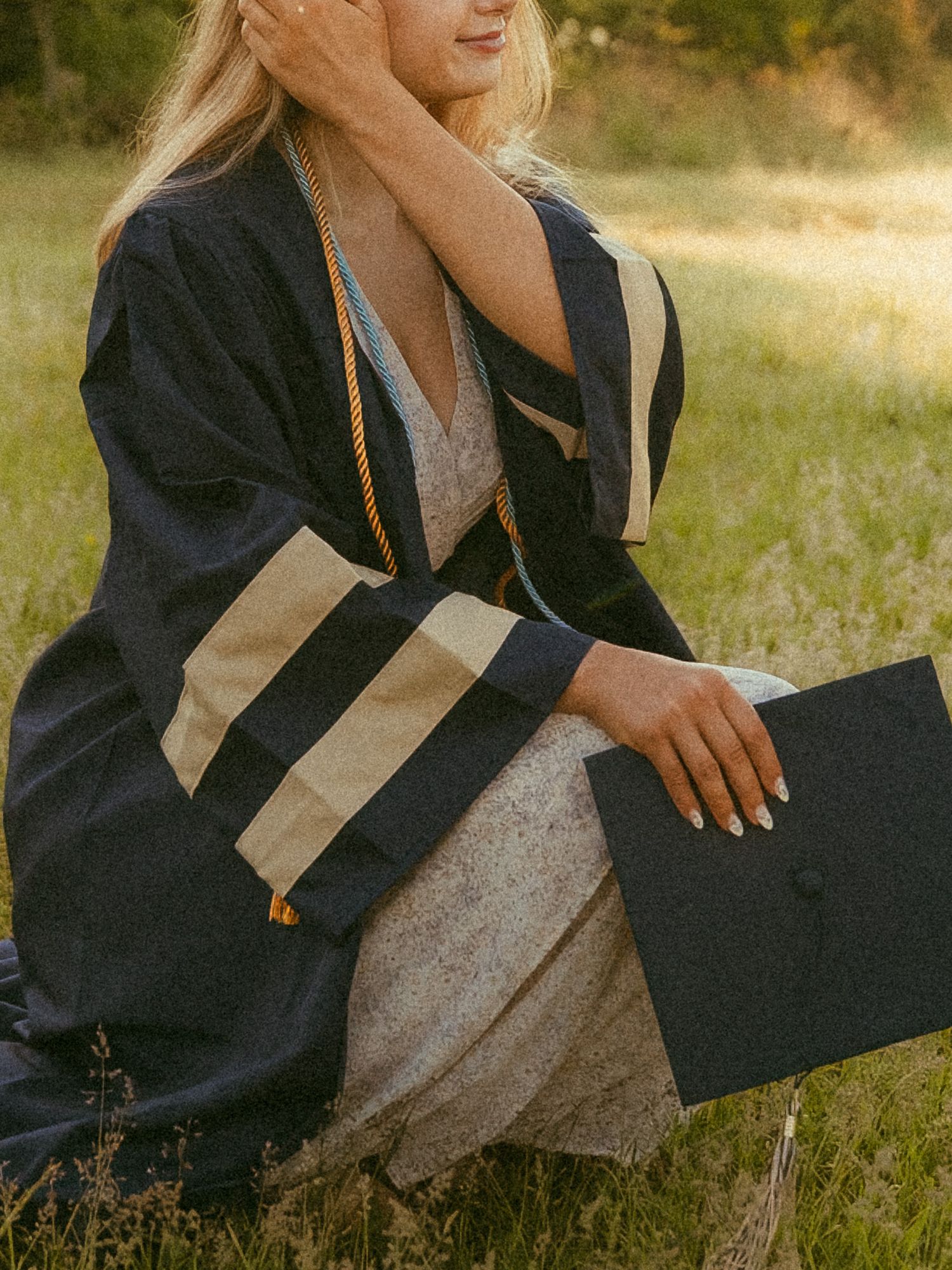 A graduate in navy blue cap and gown sits outdoors in a sunlit field, holding her graduation cap.