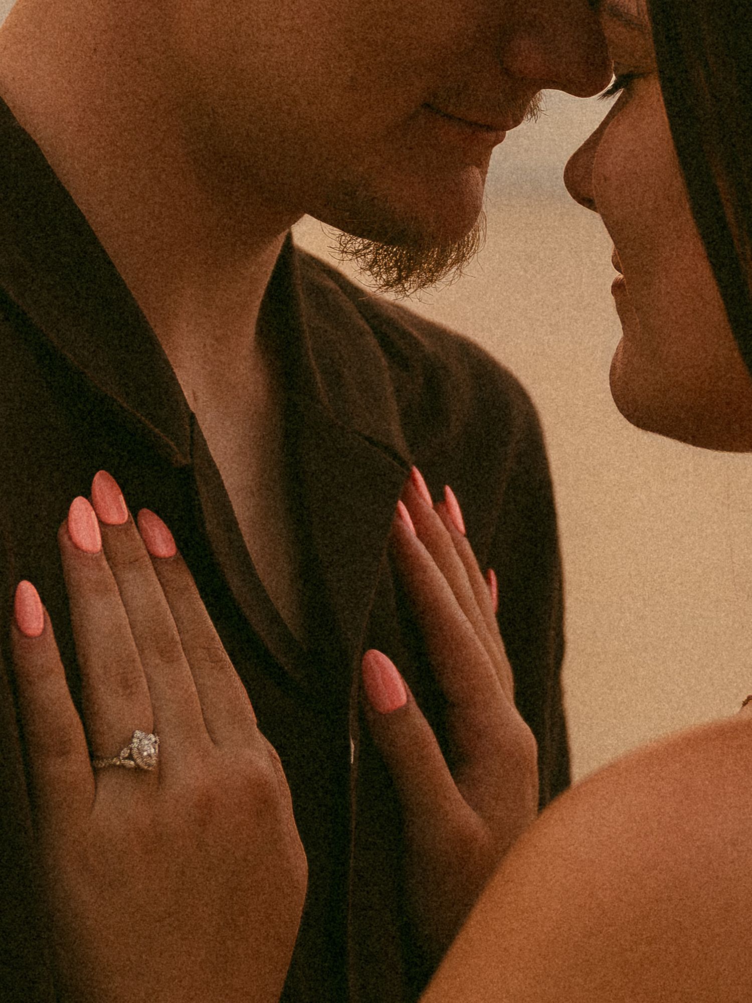 Couple sharing intimate moment with woman displaying diamond engagement ring and coral pink nails on man's chest.