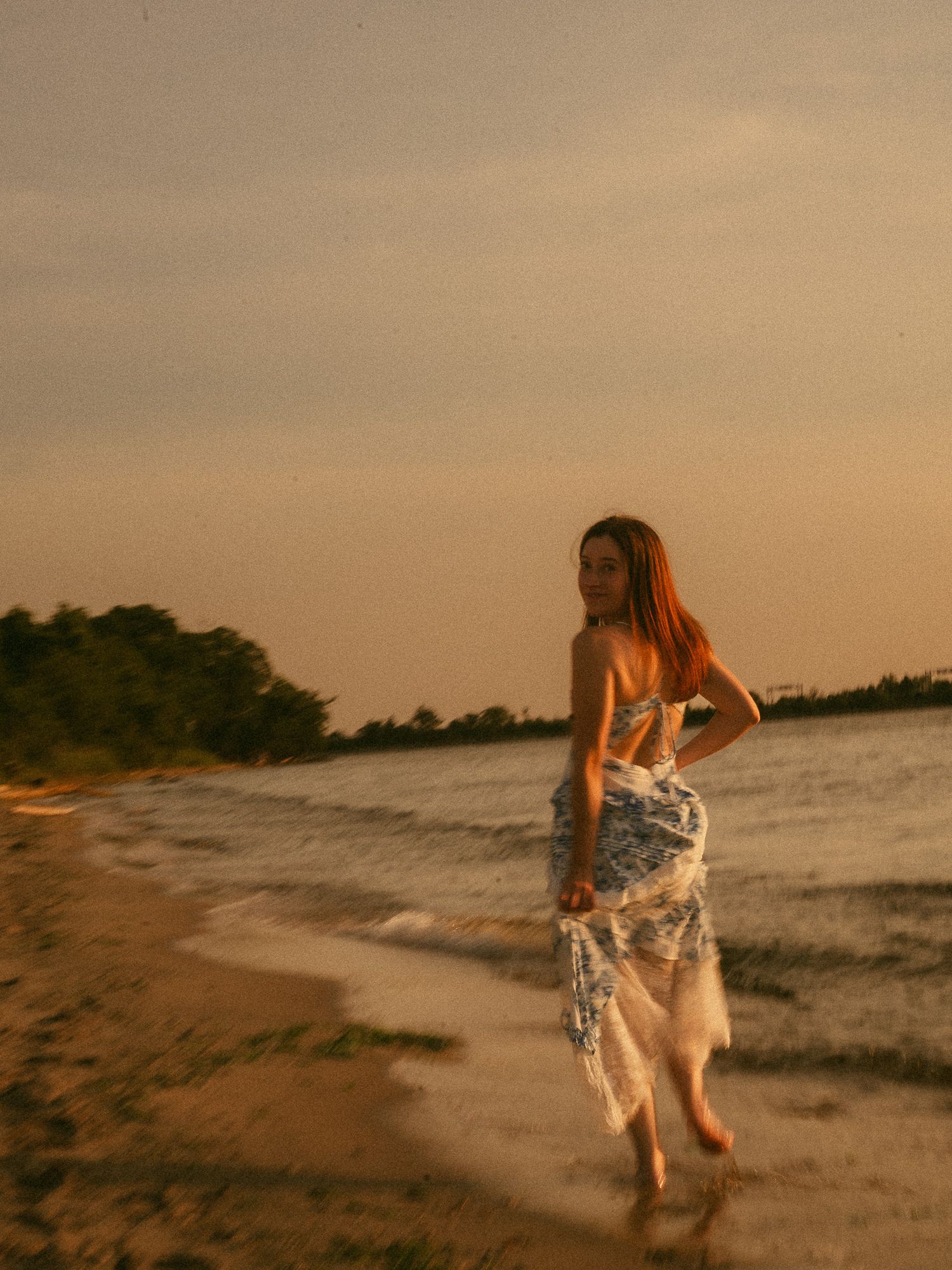 A woman in a blue floral dress walks along a sandy beach at sunset, glancing back over her shoulder.