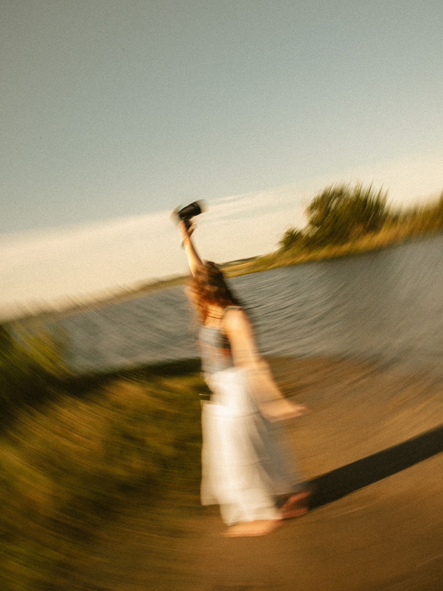A blurred motion photo of a woman in a white dress raising a camera above her head outdoors at golden hour.