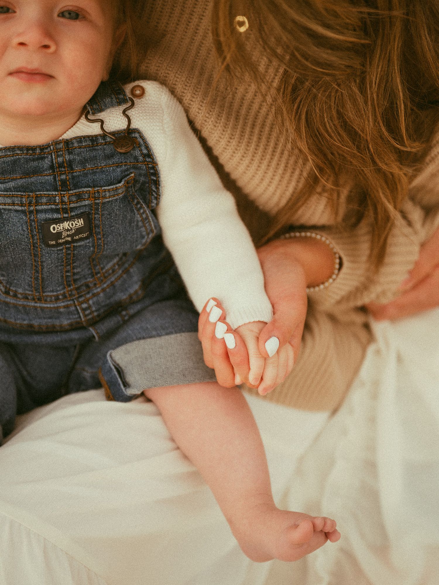 A mother gently holds her baby's hand, dressed in denim overalls, in a warm, soft-toned portrait.