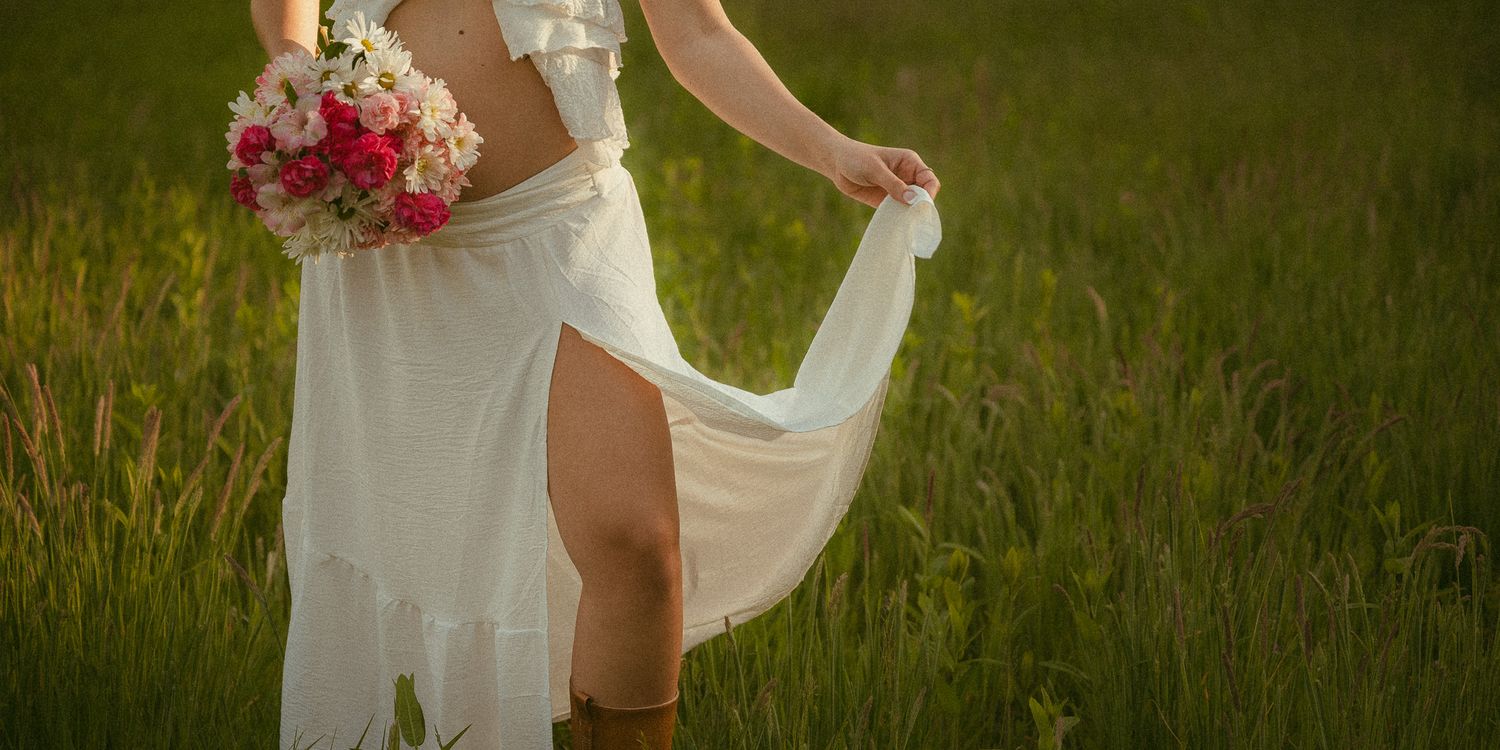 Bride in white dress with slit and cowboy boots holding pink bouquet in green field.