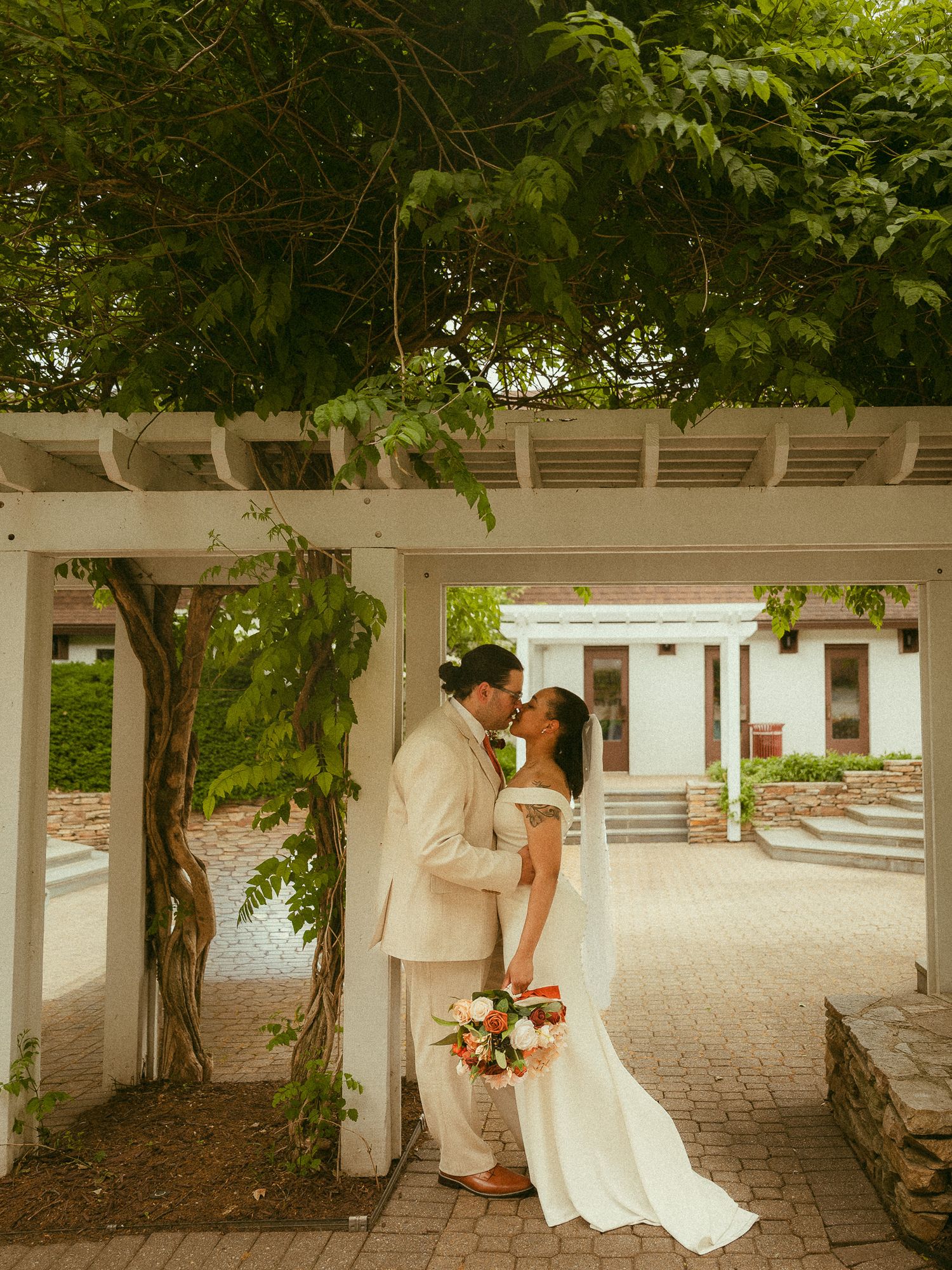 Bride and groom sharing a kiss under a white pergola with lush greenery, bride holding a bouquet on a brick patio.
